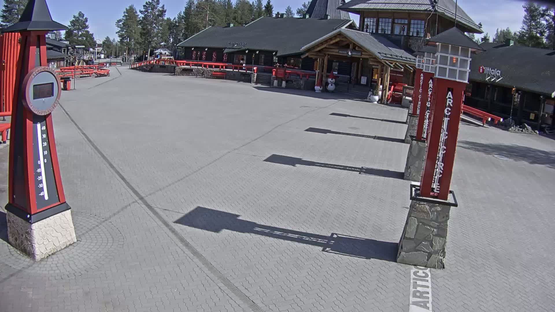A paved plaza in sunny weather with several tall, red, rectangular signs and a large thermometer-like structure.