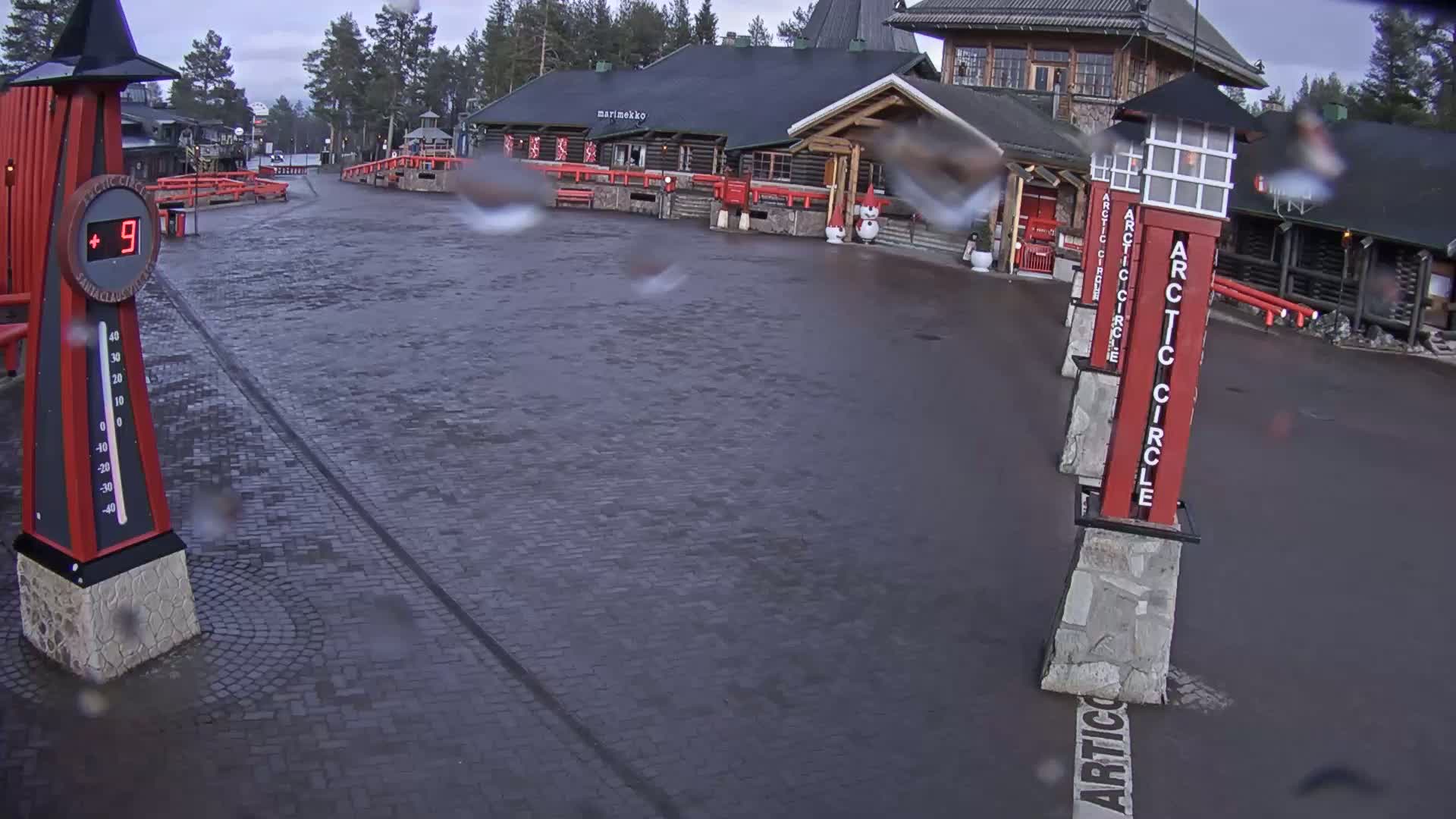 A wet, brick plaza with red Arctic Circle signposts and a thermometer showing 9 degrees, in front of a log cabin building.