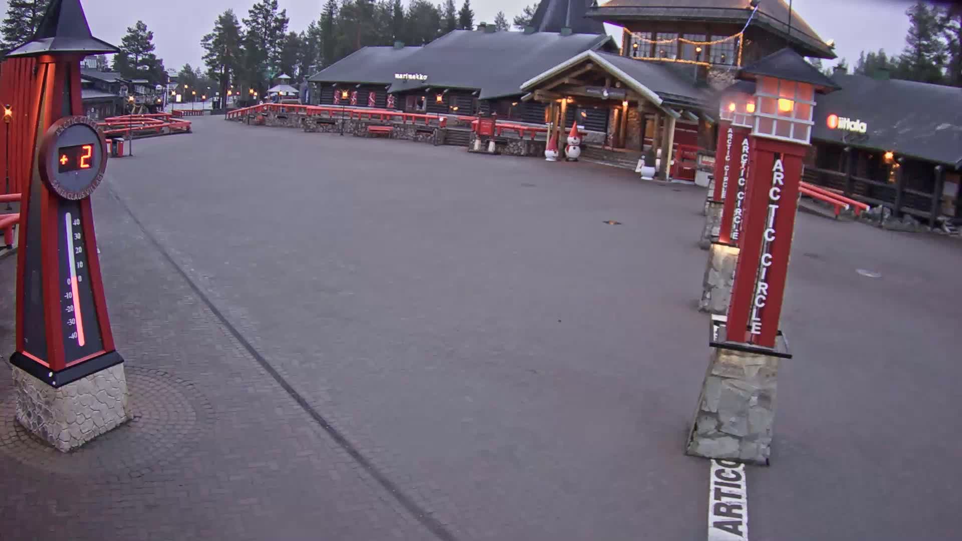 A paved plaza with red wooden buildings and illuminated signs under an overcast sky, showing a temperature of 2 degrees.