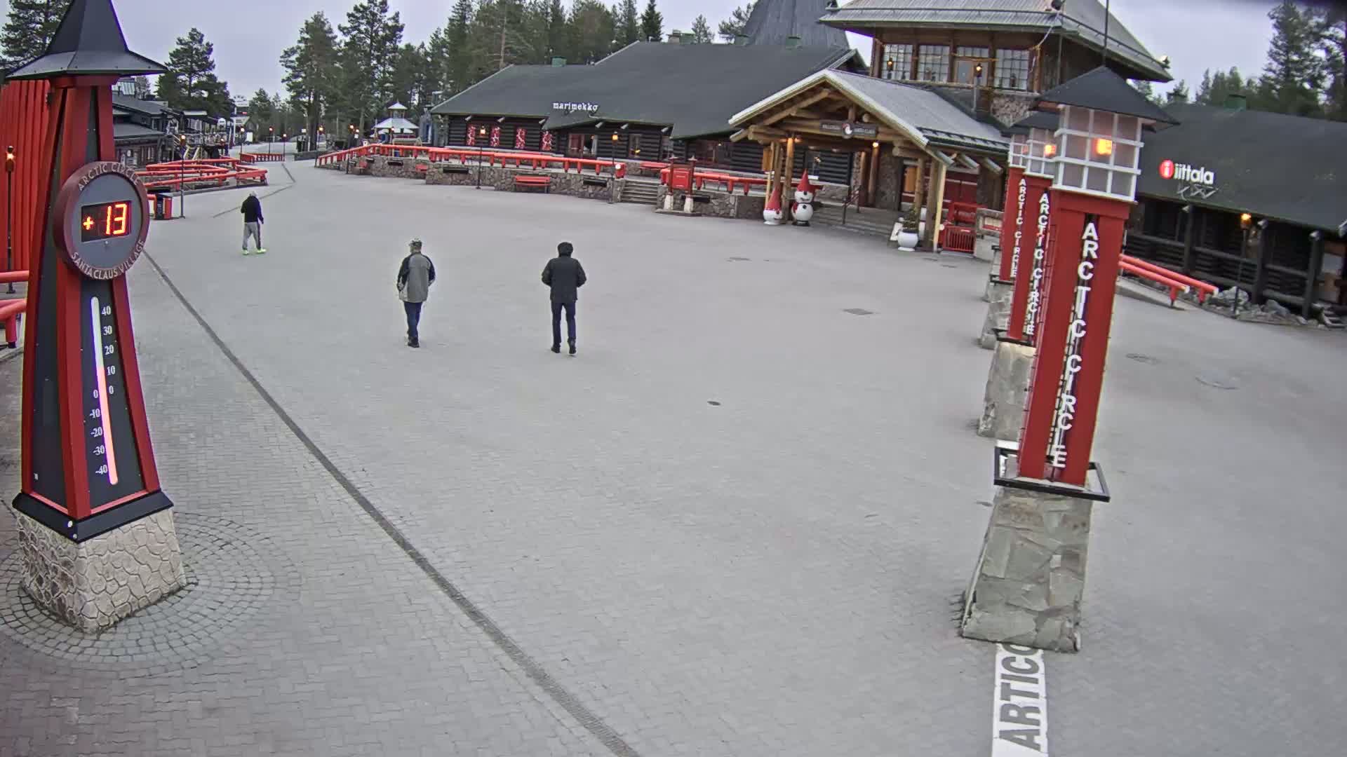 A paved plaza with a few pedestrians, log buildings in the background, and a thermometer showing thirteen degrees, under an overcast sky.