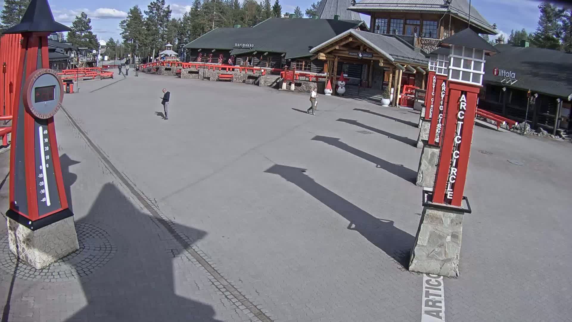 A paved plaza with several people walking, a large thermometer, and tall red and gray pillars on a sunny day.