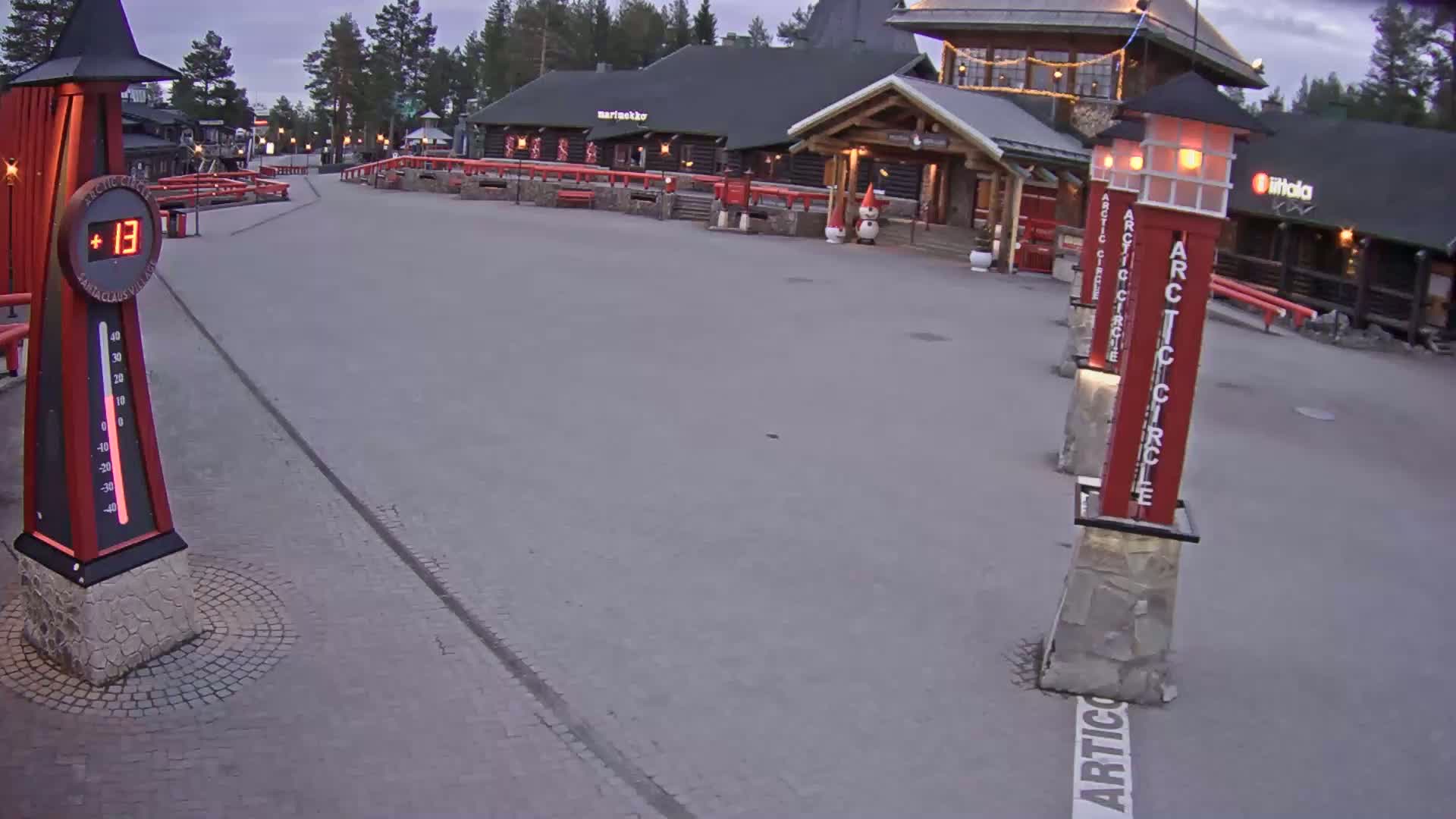 A paved plaza with several red and stone pillars, a large thermometer showing 13 degrees, and buildings in the background under a twilight sky.