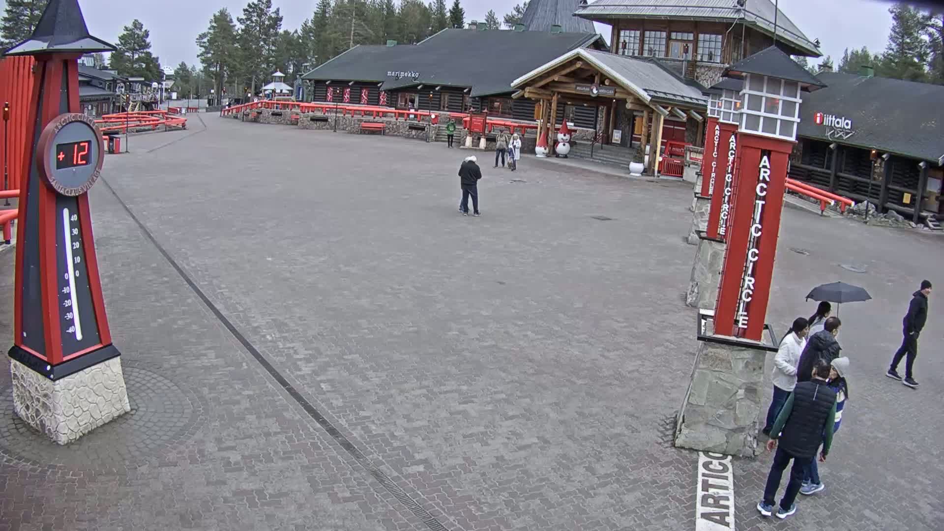 A paved plaza with several people walking around under an overcast sky, featuring red Arctic Circle signs and a large thermometer displaying 12 degrees.