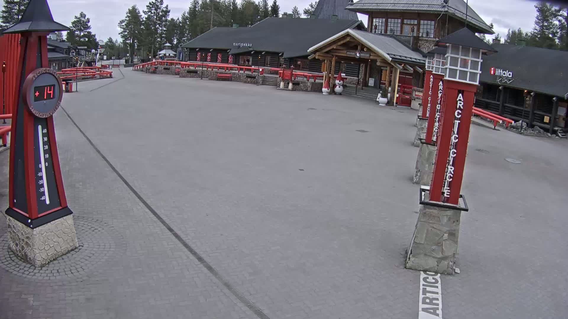 A paved plaza with several red Arctic Circle markers and a large thermometer showing a temperature of 14 degrees, situated in front of a log cabin-style building under an overcast sky.