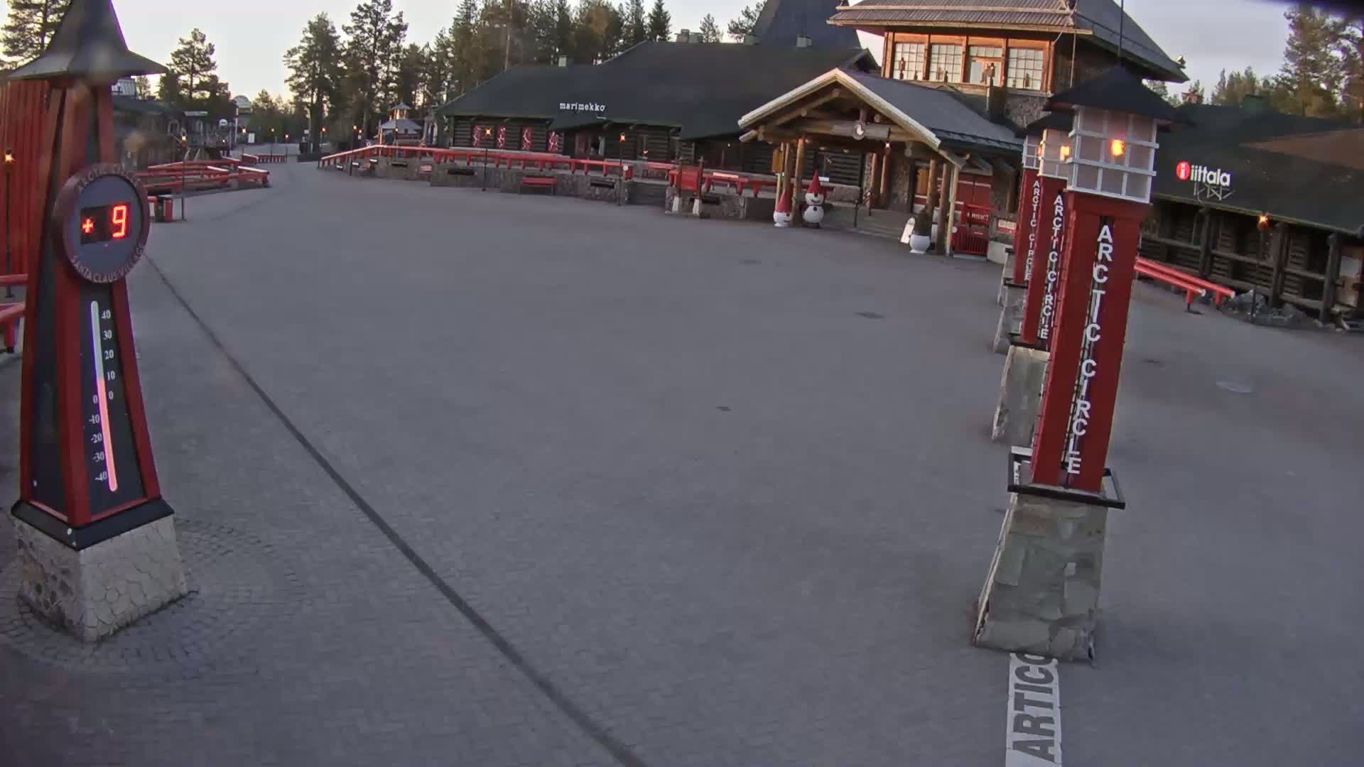 A paved plaza shows a thermometer reading plus nine degrees, several red posts with "Arctic Circle" written on them, and a log cabin-style building in the background under a twilight sky.