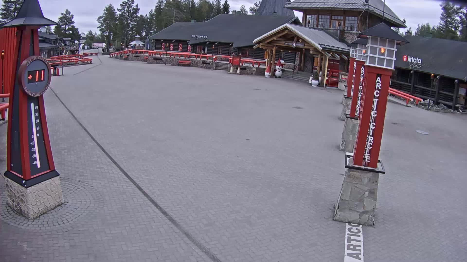 An overcast day shows an empty paved plaza in front of a log building, with several tall red pillars and a large thermometer displaying a temperature above freezing.