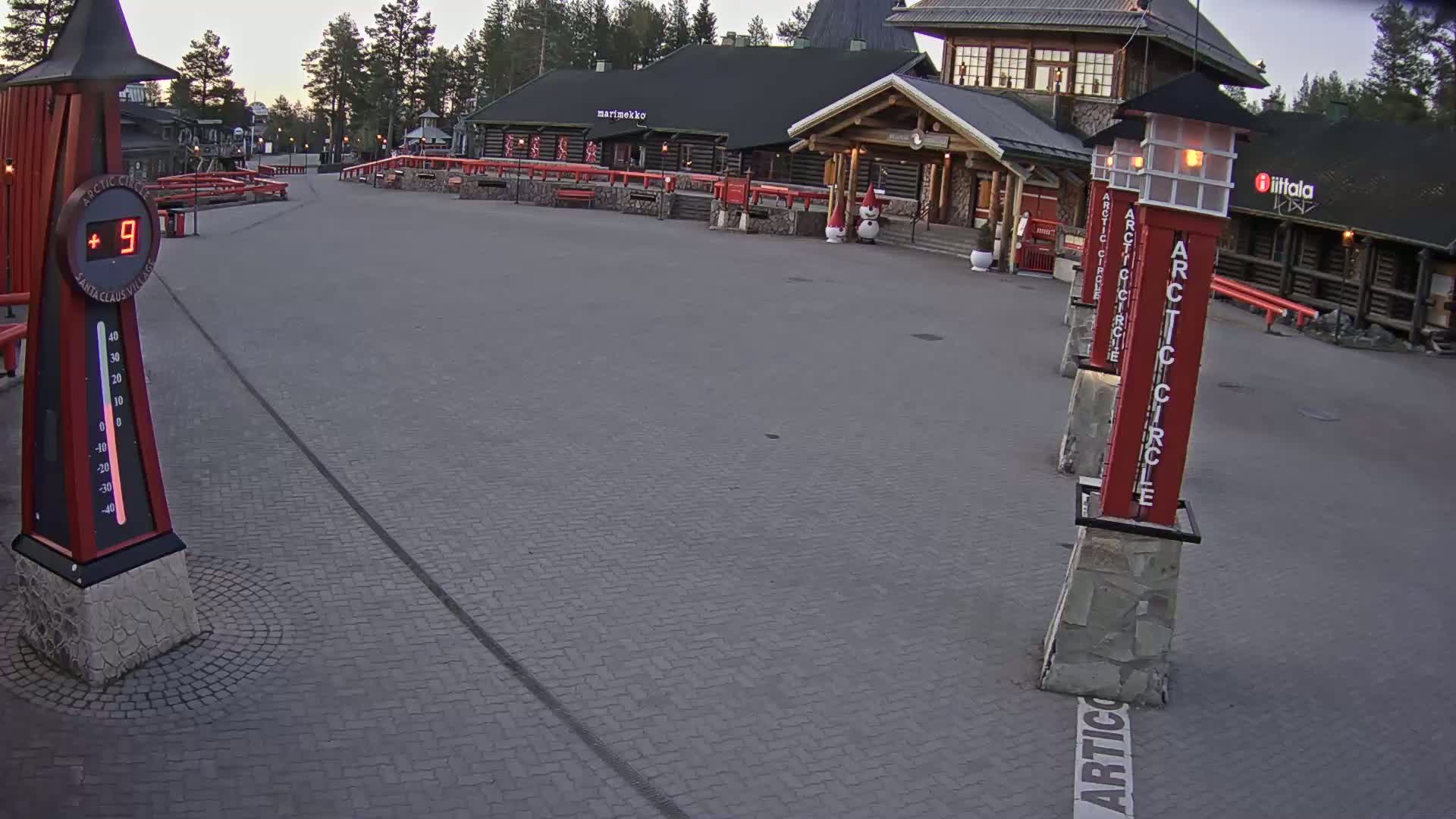 A paved plaza with several buildings, a thermometer showing 9 degrees, and tall, red pillars marking the Arctic Circle under a twilight sky.