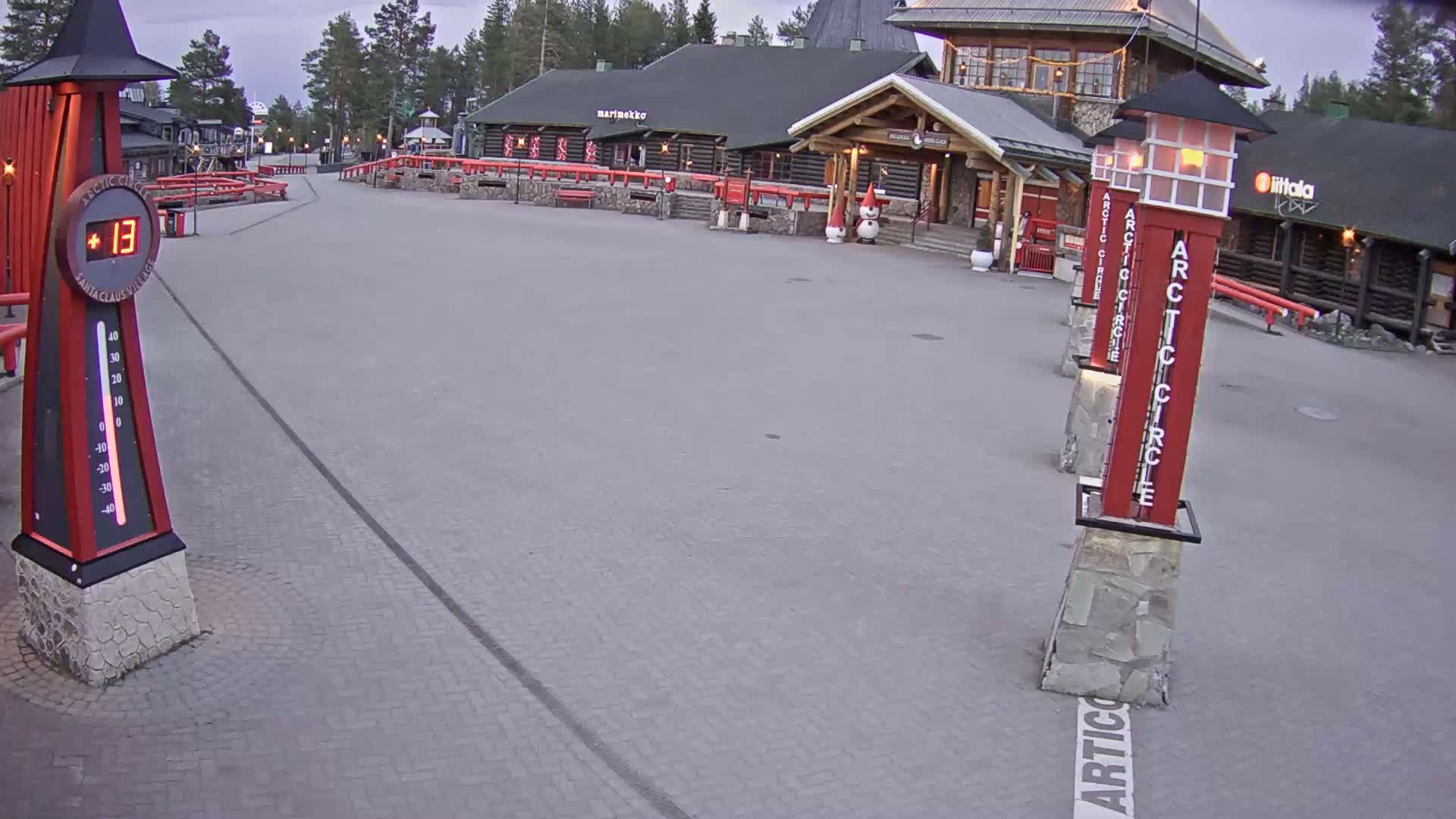 An overcast outdoor scene shows a paved plaza with several buildings and tall red posts that mark the Arctic Circle, along with a thermometer displaying a temperature of 13 degrees.