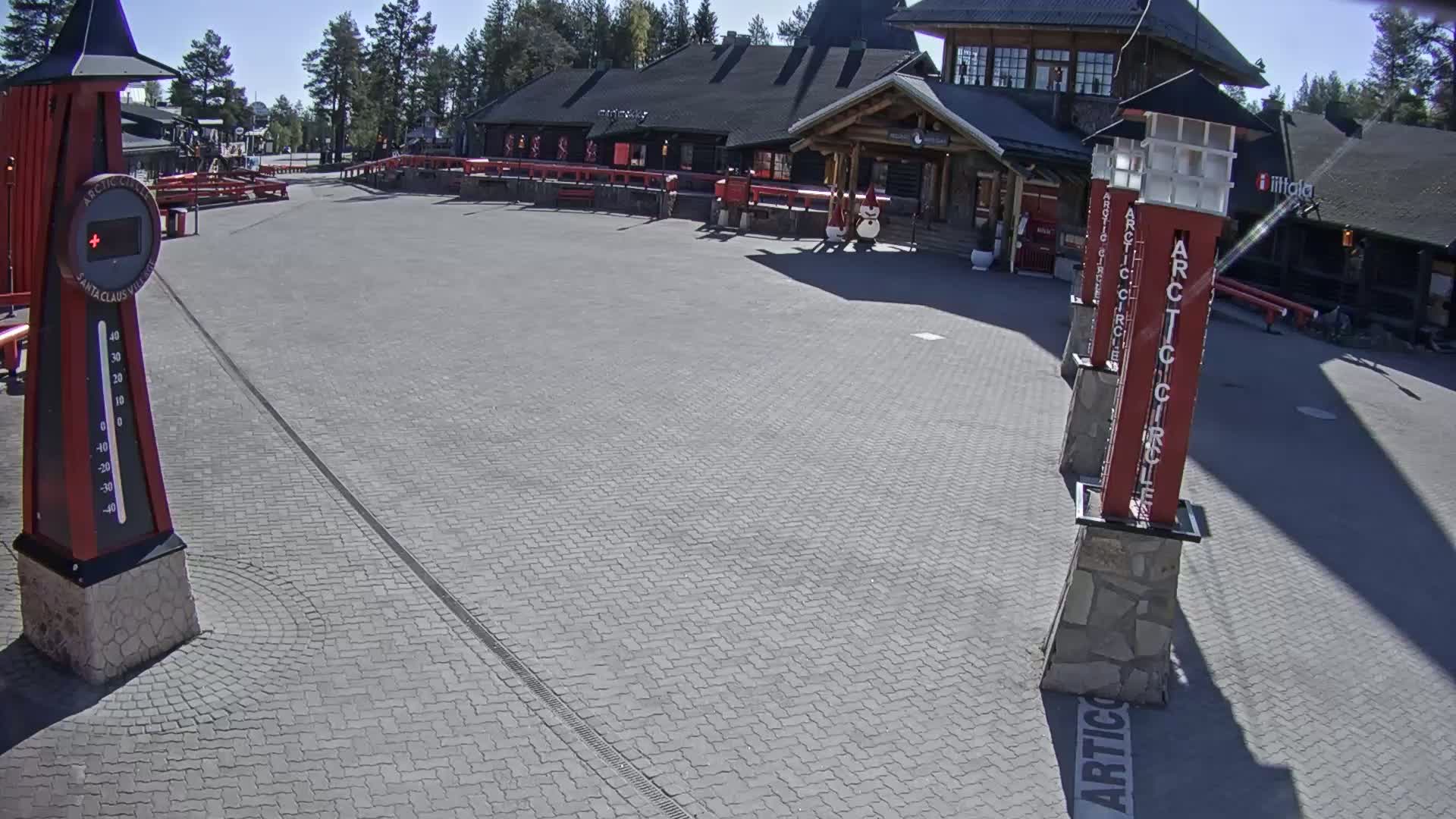 A paved plaza, under sunny skies, features a large building and several tall, red and white posts.