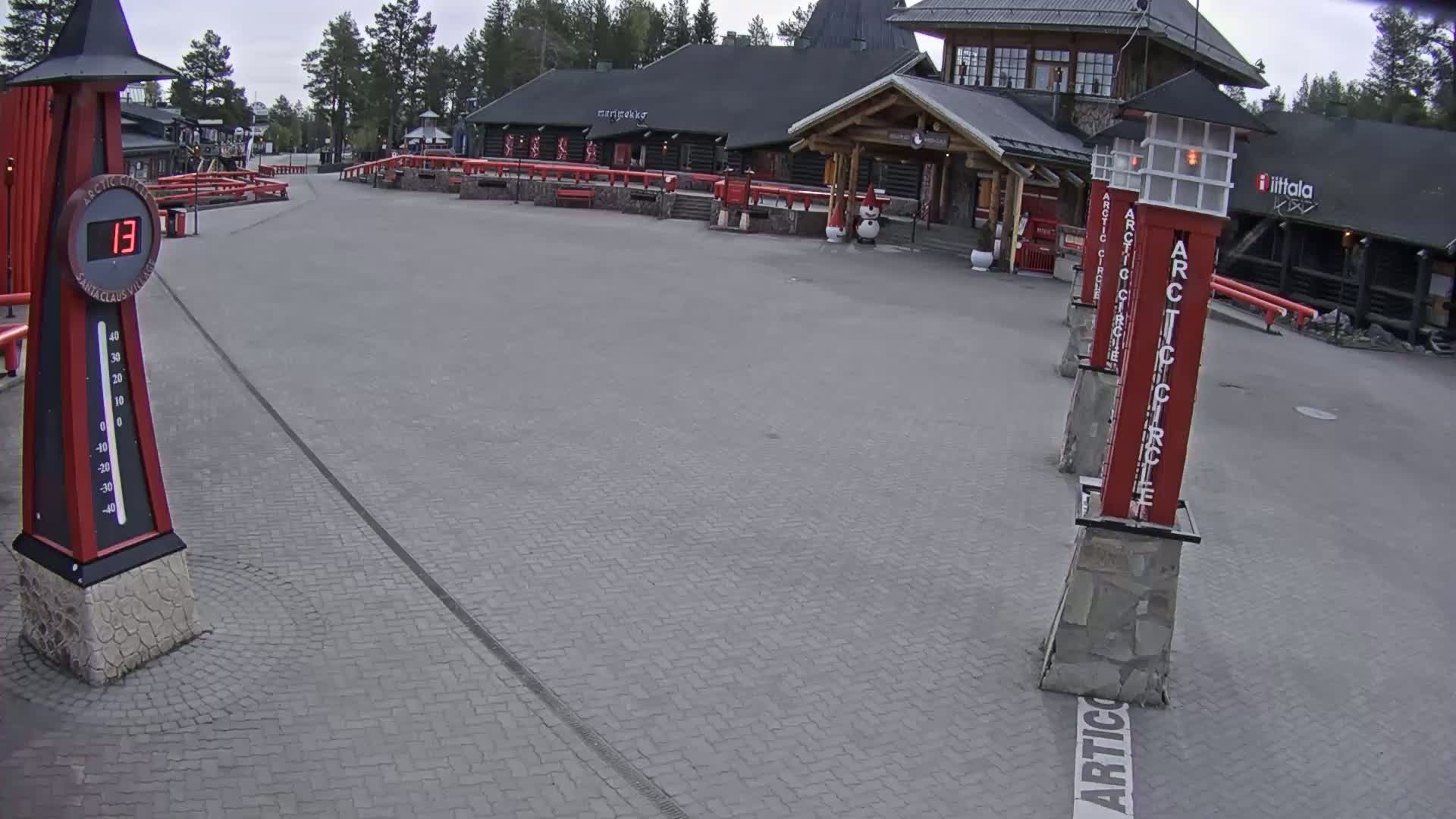 A brick paved area in front of a log cabin-style building on an overcast day, with a thermometer showing a temperature of 13 degrees and several tall red posts with white lettering.