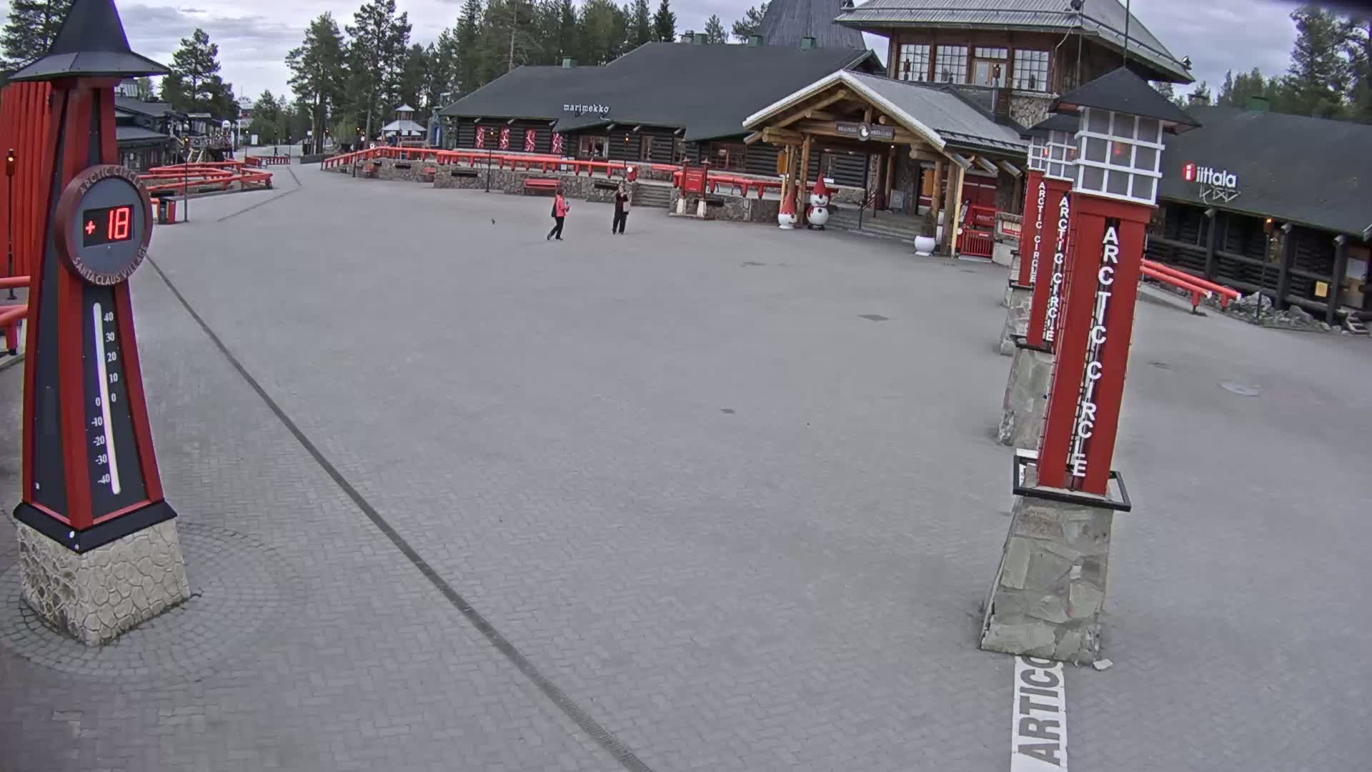A paved plaza with several people and log buildings, under an overcast sky, shows a thermometer reading 18 degrees.
