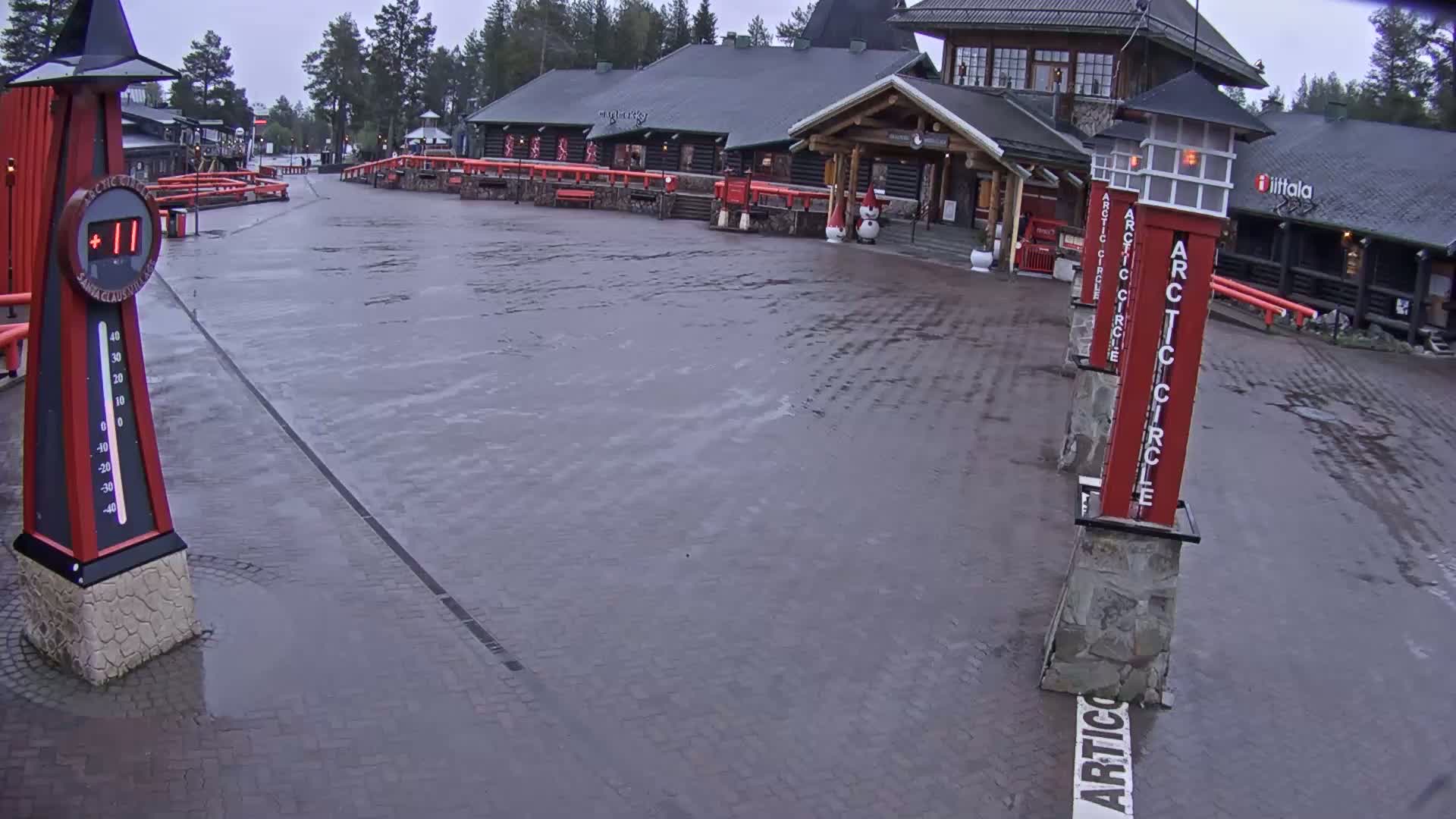 A wet, paved plaza in front of a log cabin building with several red pillars displaying the words "Arctic Circle," under an overcast sky.