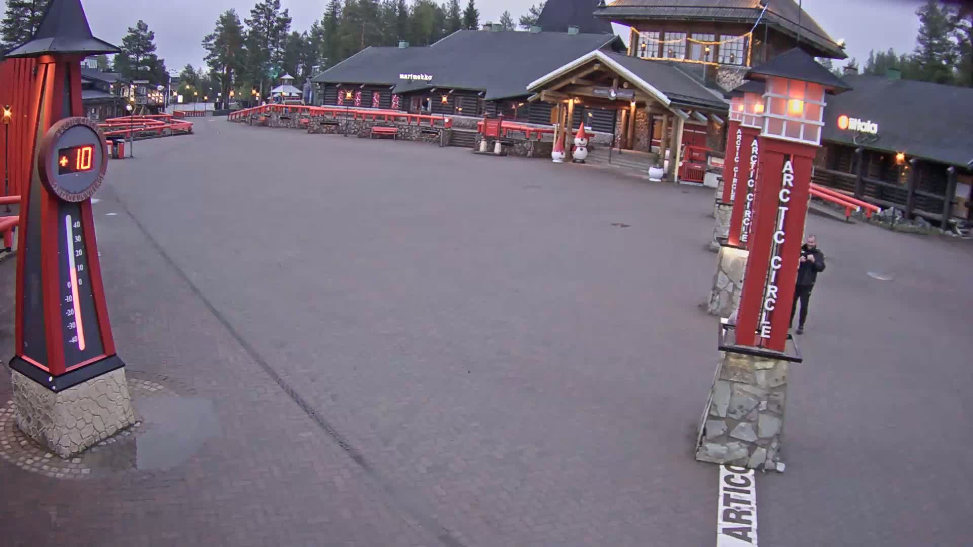 A paved area outside of several log cabins under an overcast sky shows a person standing near tall red posts marked "Arctic Circle" and a large thermometer indicating a temperature of 10 degrees.