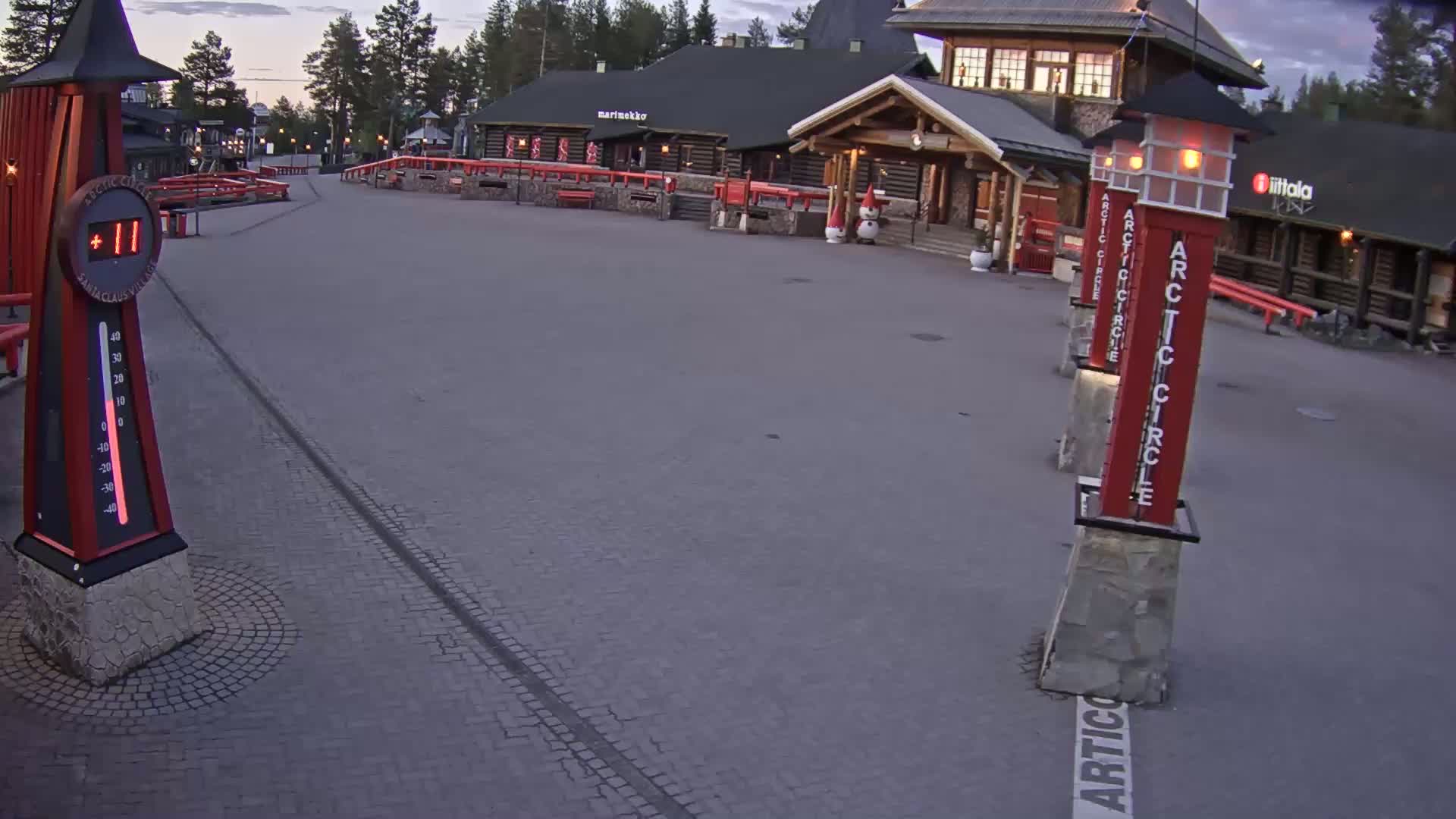A paved plaza with wooden buildings in the background, a large thermometer displaying eleven degrees, and several tall red Arctic Circle markers under a twilight sky.