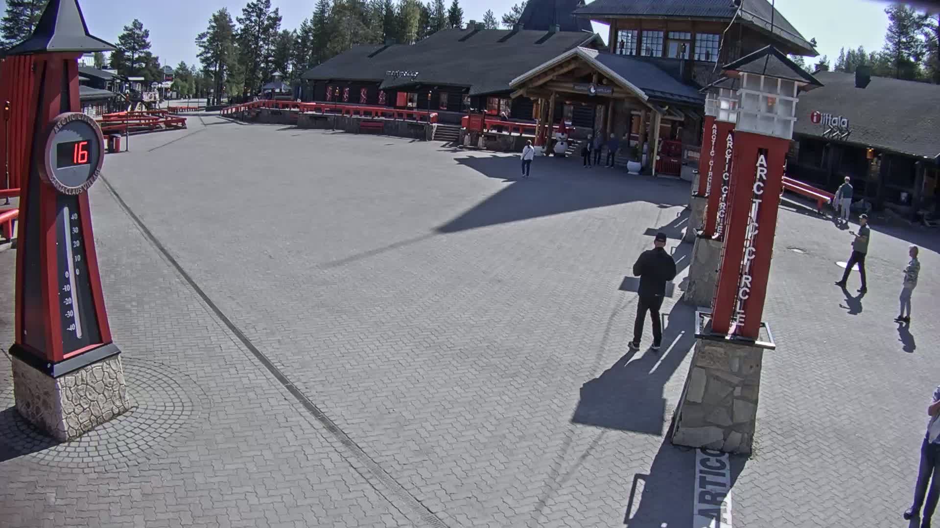 A paved plaza in front of a log building on a sunny day, with several people walking around.