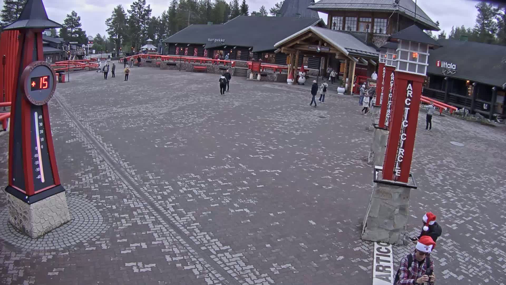 A brick plaza with several people walking around, under an overcast sky, features tall red and white Arctic Circle markers and a large thermometer showing a temperature of 15 degrees.