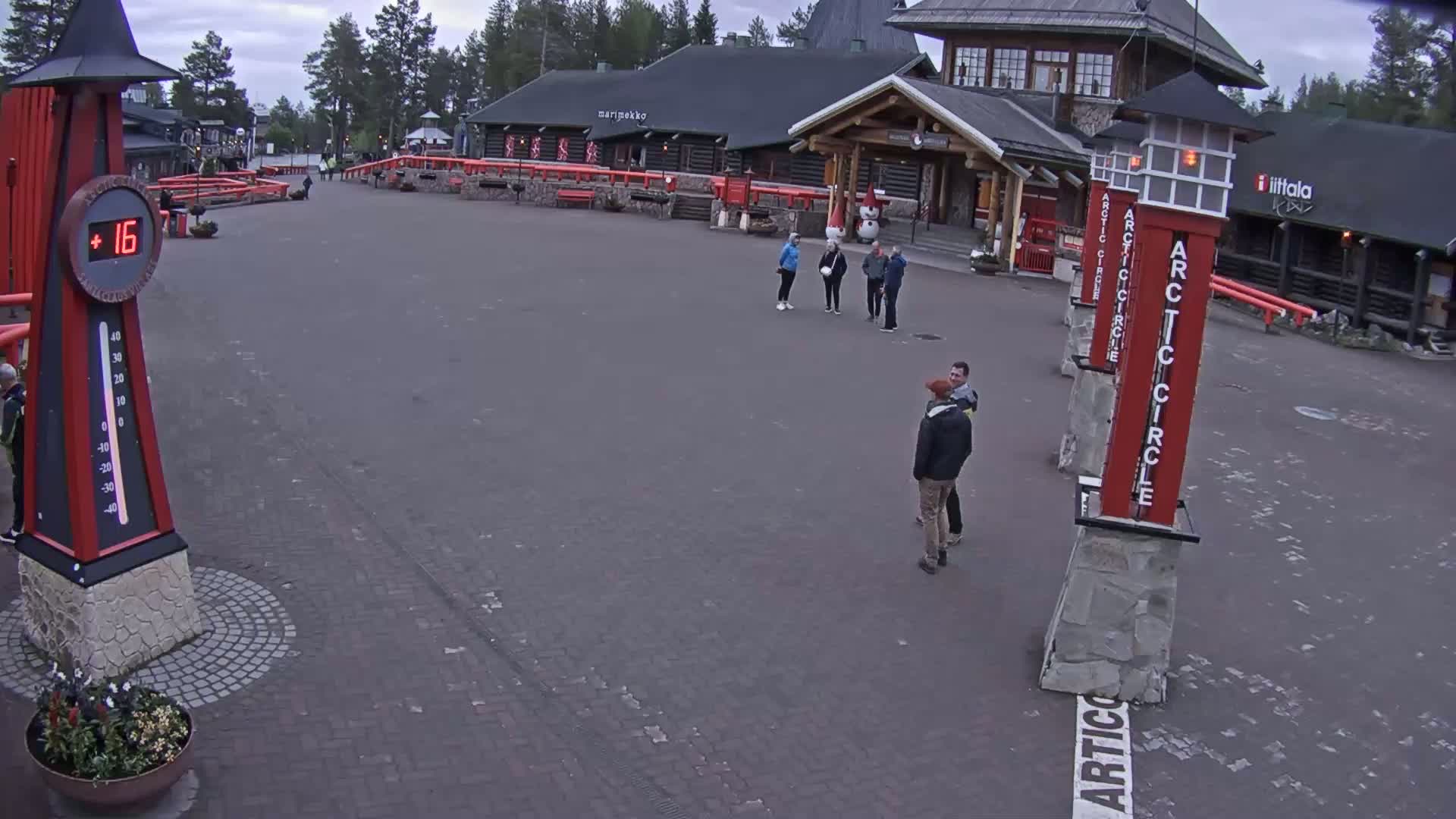 A paved plaza with several people, a large thermometer showing 16 degrees, and several tall, red and gray Arctic Circle markers under an overcast sky.