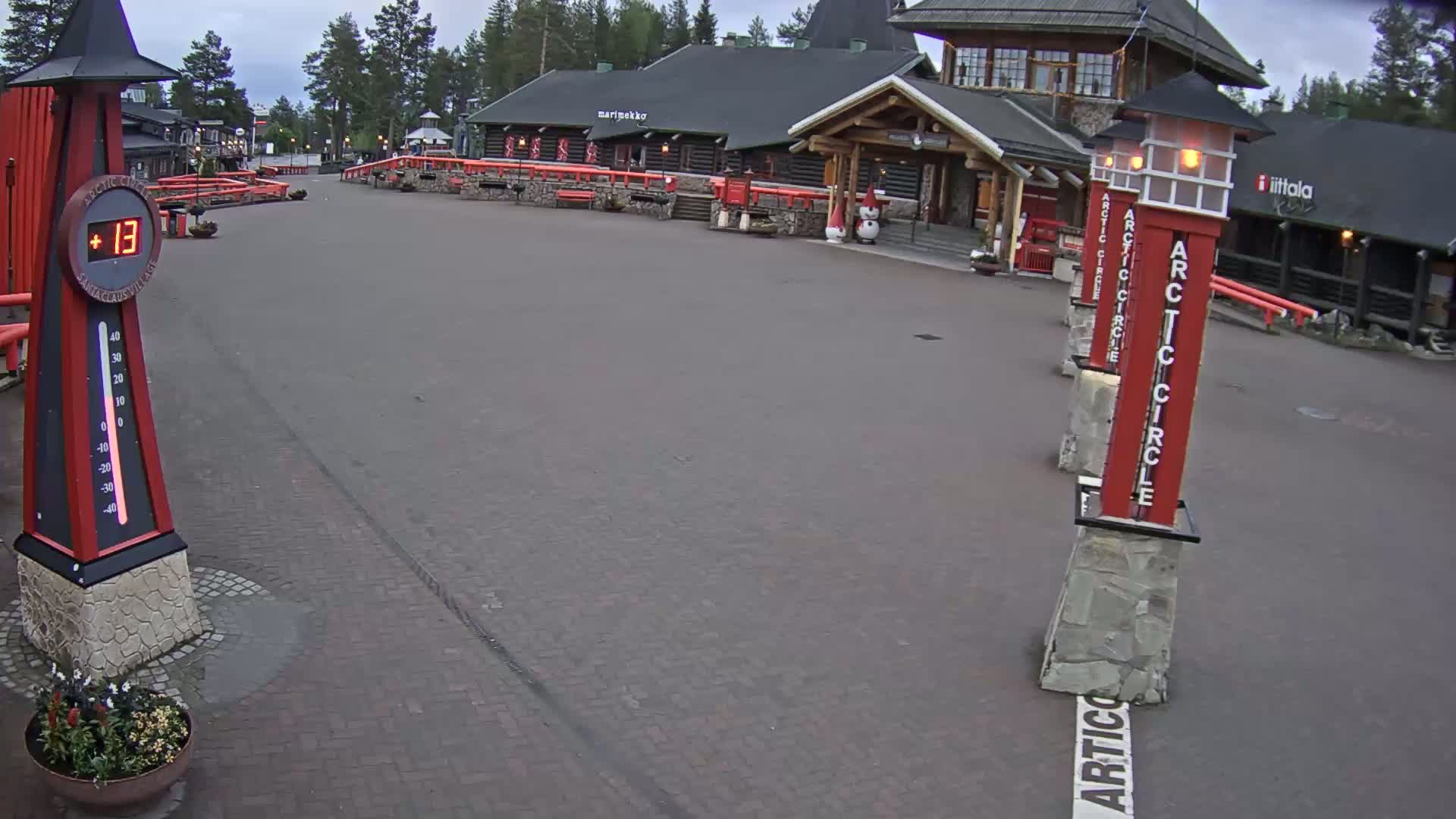 A paved plaza with a large thermometer displaying thirteen degrees, several red and white Arctic Circle posts, and a log cabin-style building in the background under an overcast sky.