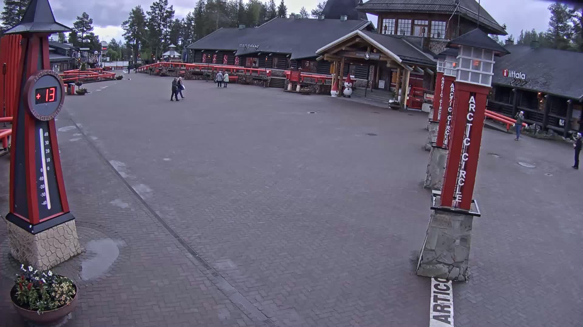 A brick paved plaza with several people walking near buildings under an overcast sky, a thermometer showing 13 degrees, and several red pillars marking the Arctic Circle.
