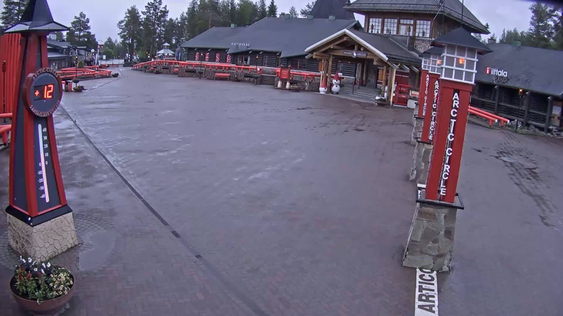A wet, brick plaza sits in front of a cluster of log buildings under an overcast sky, with several tall red signs marking the Arctic Circle.
