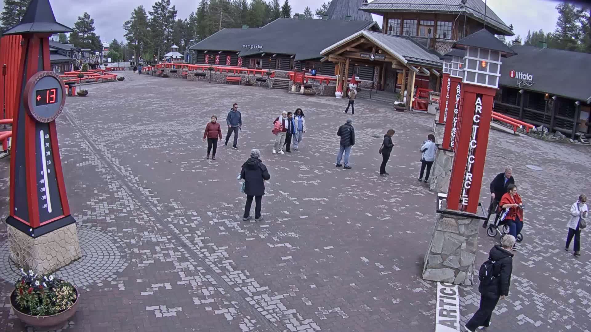 A paved plaza with several people walking around under an overcast sky, near buildings with log cabin architecture.