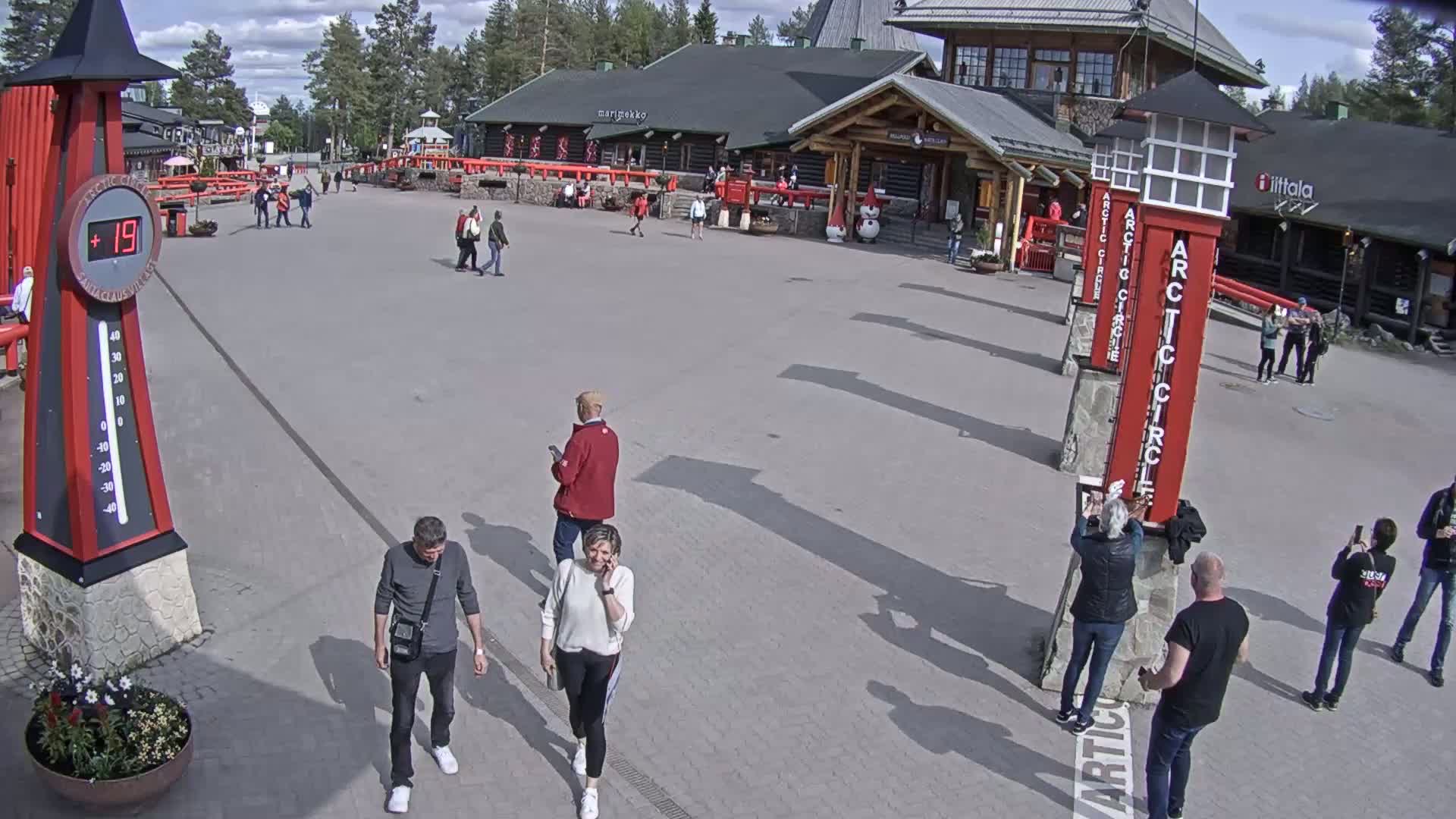A paved plaza with several people walking around, near buildings under a partly sunny sky.