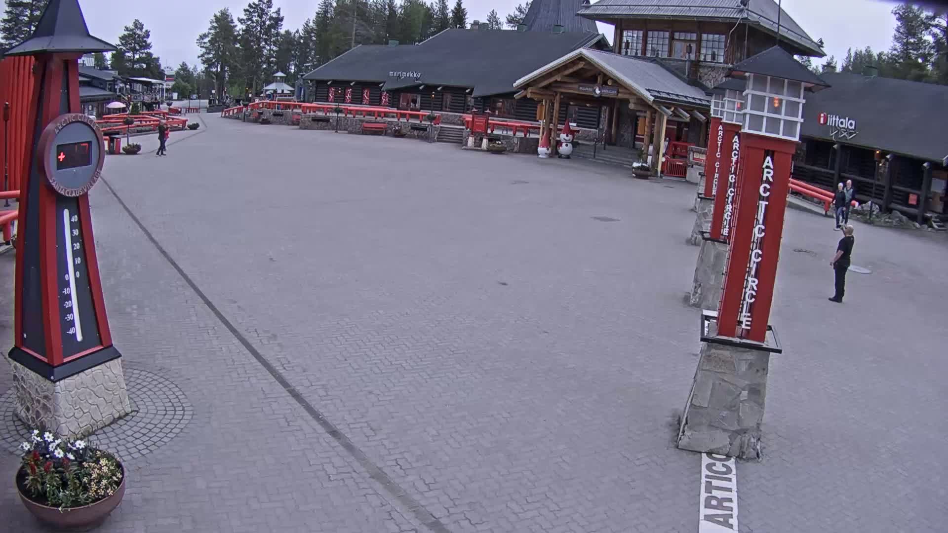 A paved plaza with several buildings and a large thermometer displays a temperature, under an overcast sky.