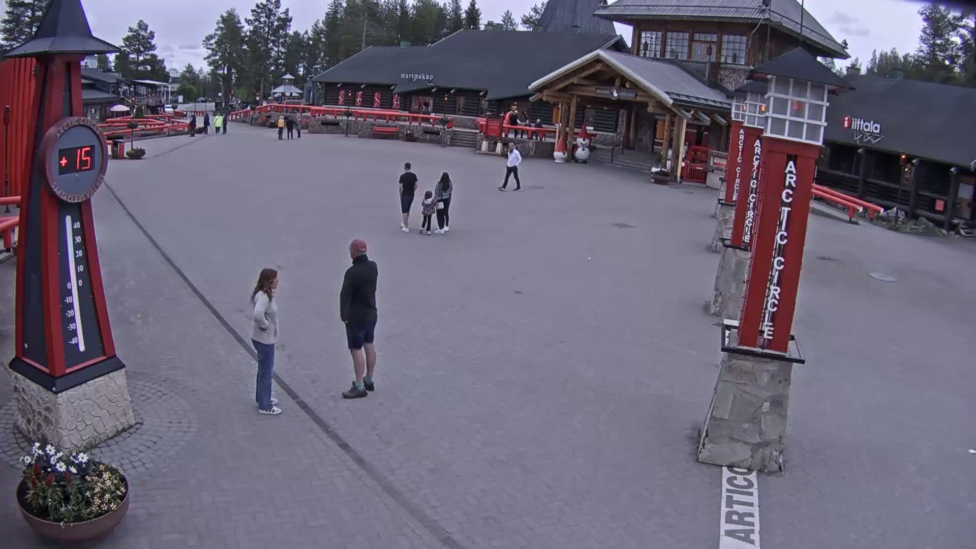 A paved plaza with several people, a thermometer showing 15 degrees, and several buildings under an overcast sky.