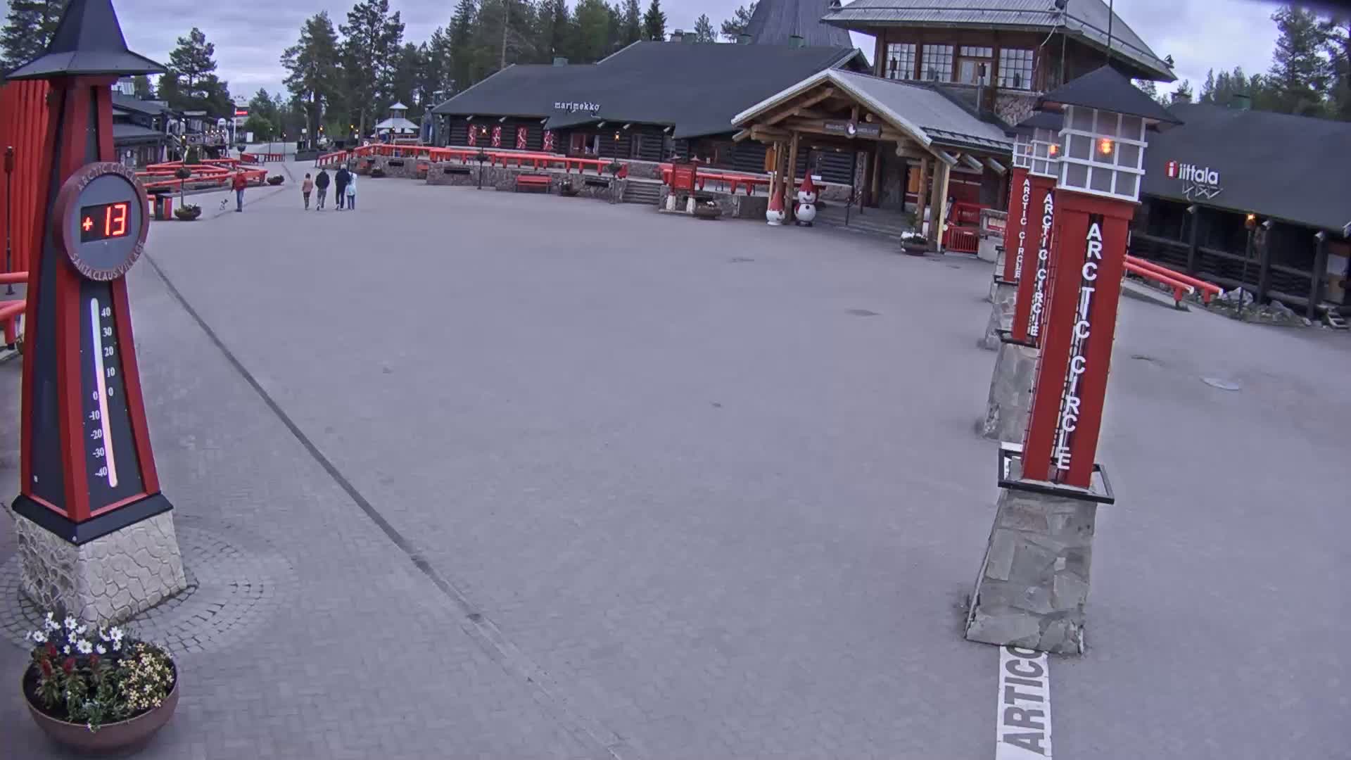 A paved plaza with several people walking near log buildings under an overcast sky, and a thermometer showing a temperature of 13 degrees.