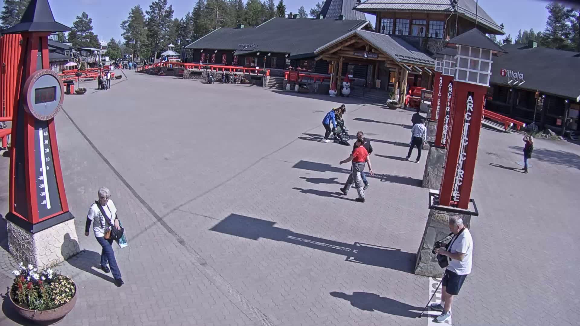 A paved plaza with several people walking, a large thermometer, and a building in the background on a sunny day.