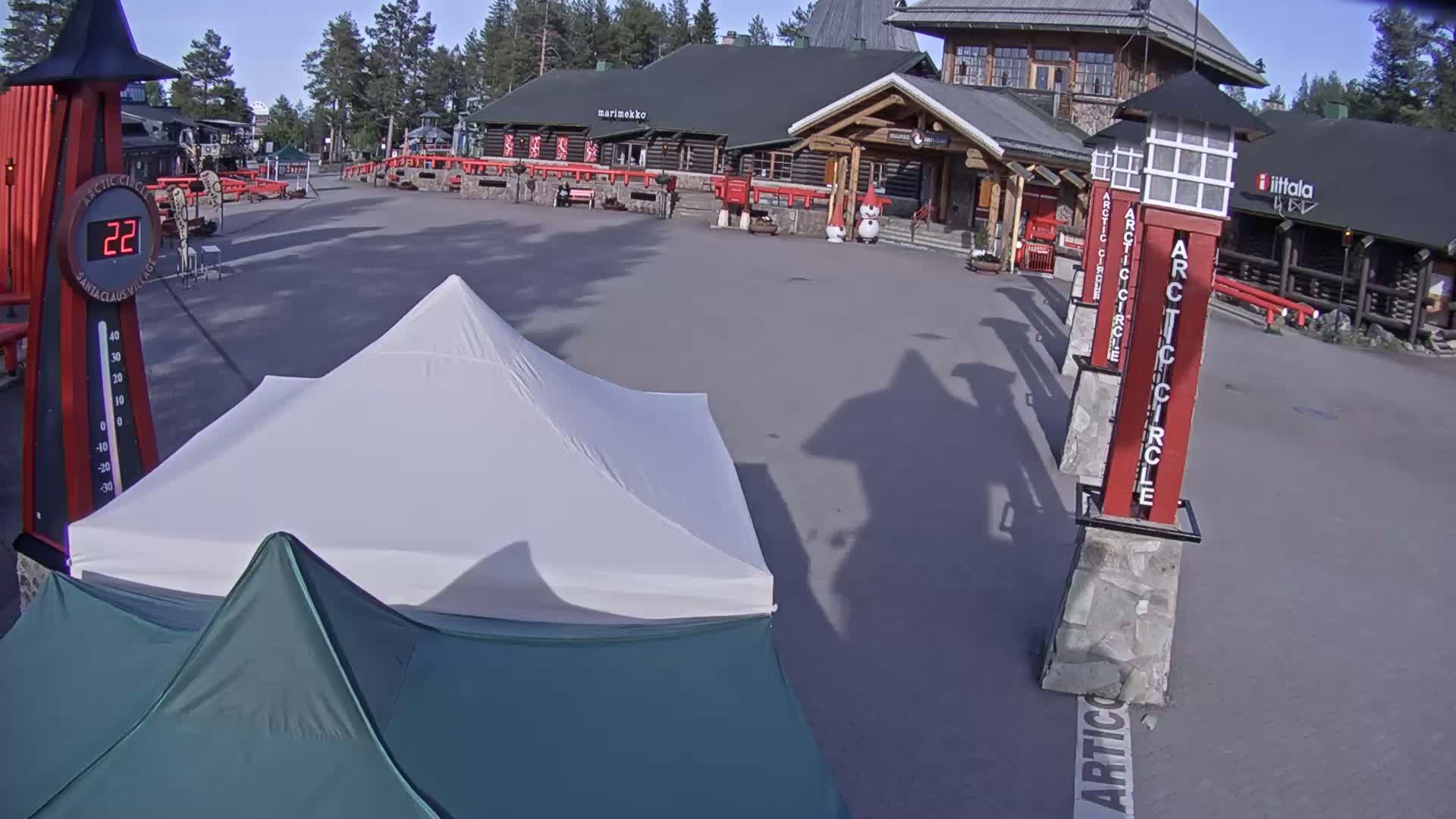 A paved plaza with a white tent and a dark-green tent, situated in front of a log cabin-style building on a sunny day.