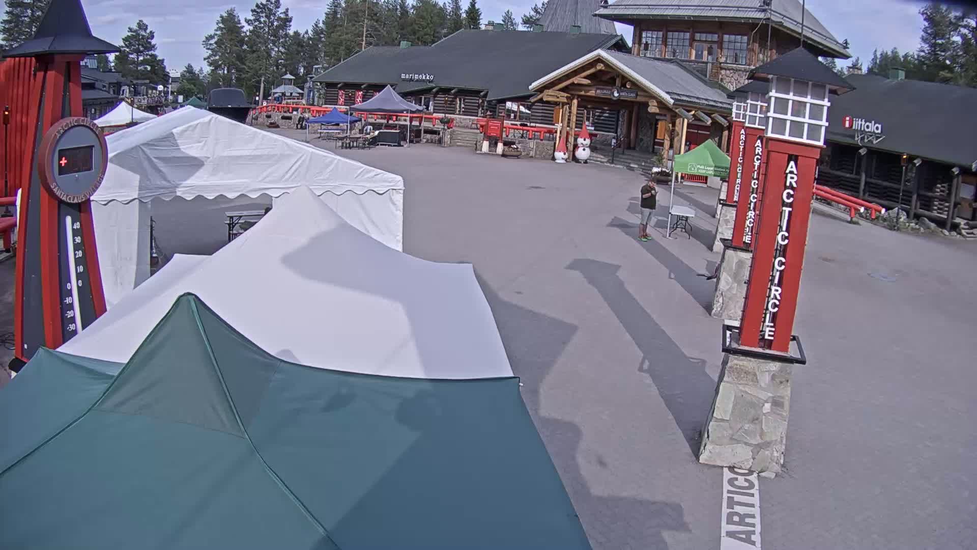 A mostly sunny outdoor scene shows a paved plaza with several tents, a log building, and tall red signs marking the Arctic Circle.