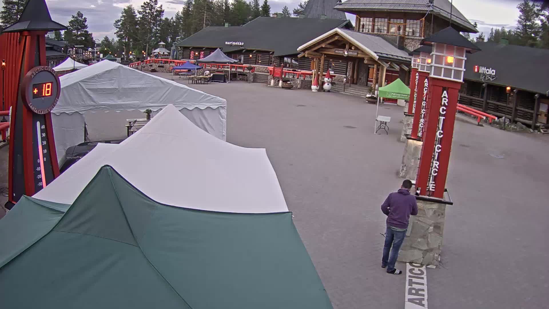 A mostly sunny outdoor scene shows a paved plaza with several tents, a log building, and tall red signs marking the Arctic Circle.