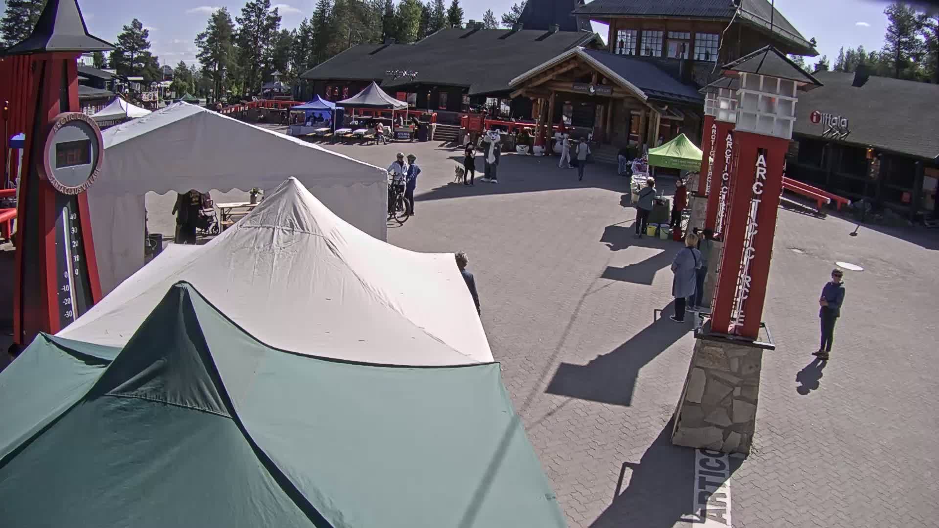 A sunny outdoor plaza with several people, market stalls under tents, and tall red pillars.