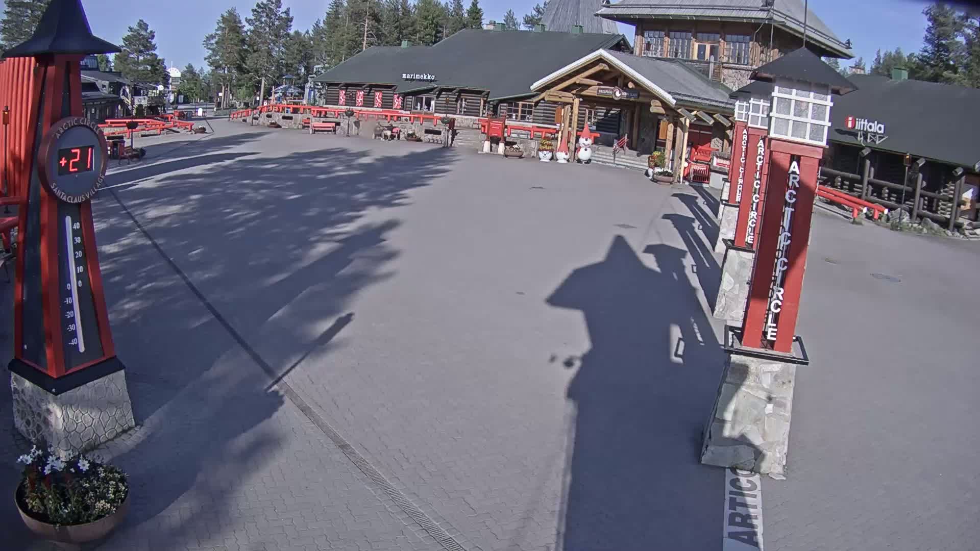 A paved plaza with several buildings, a thermometer showing 21 degrees, and tall red pillars with "Arctic Circle" written on them under a sunny sky.