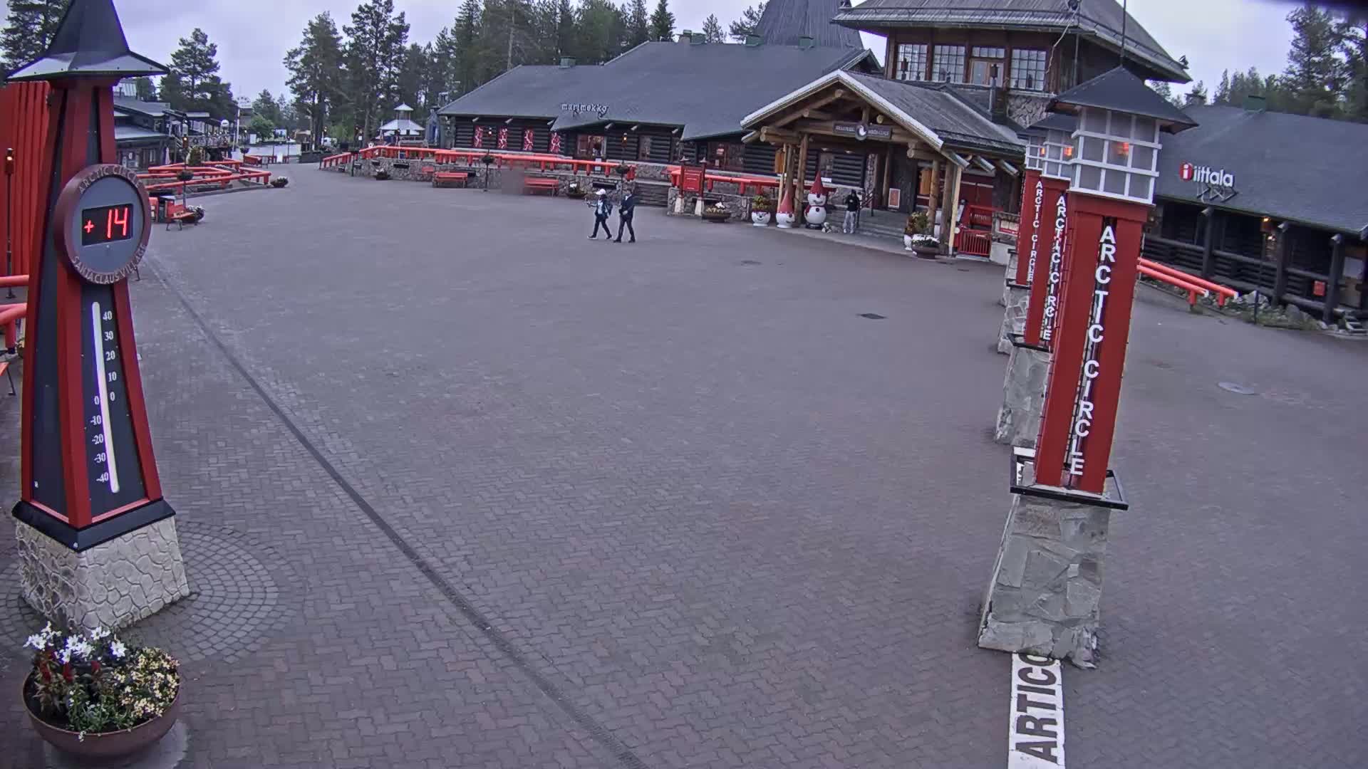 A brick plaza with a few people walking near a log cabin-style building on an overcast day, a large thermometer showing a temperature of 14 degrees, and several tall, red Arctic Circle signposts.