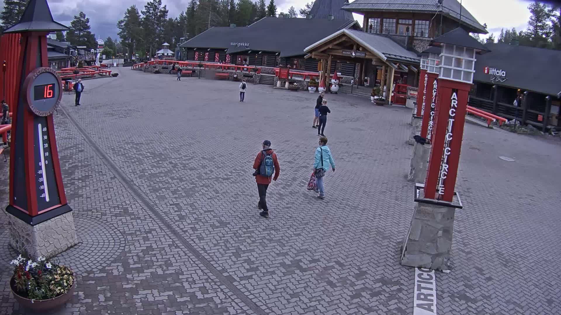 A paved plaza with several people walking near buildings under a cloudy sky, and a large thermometer indicating a temperature of 16 degrees.