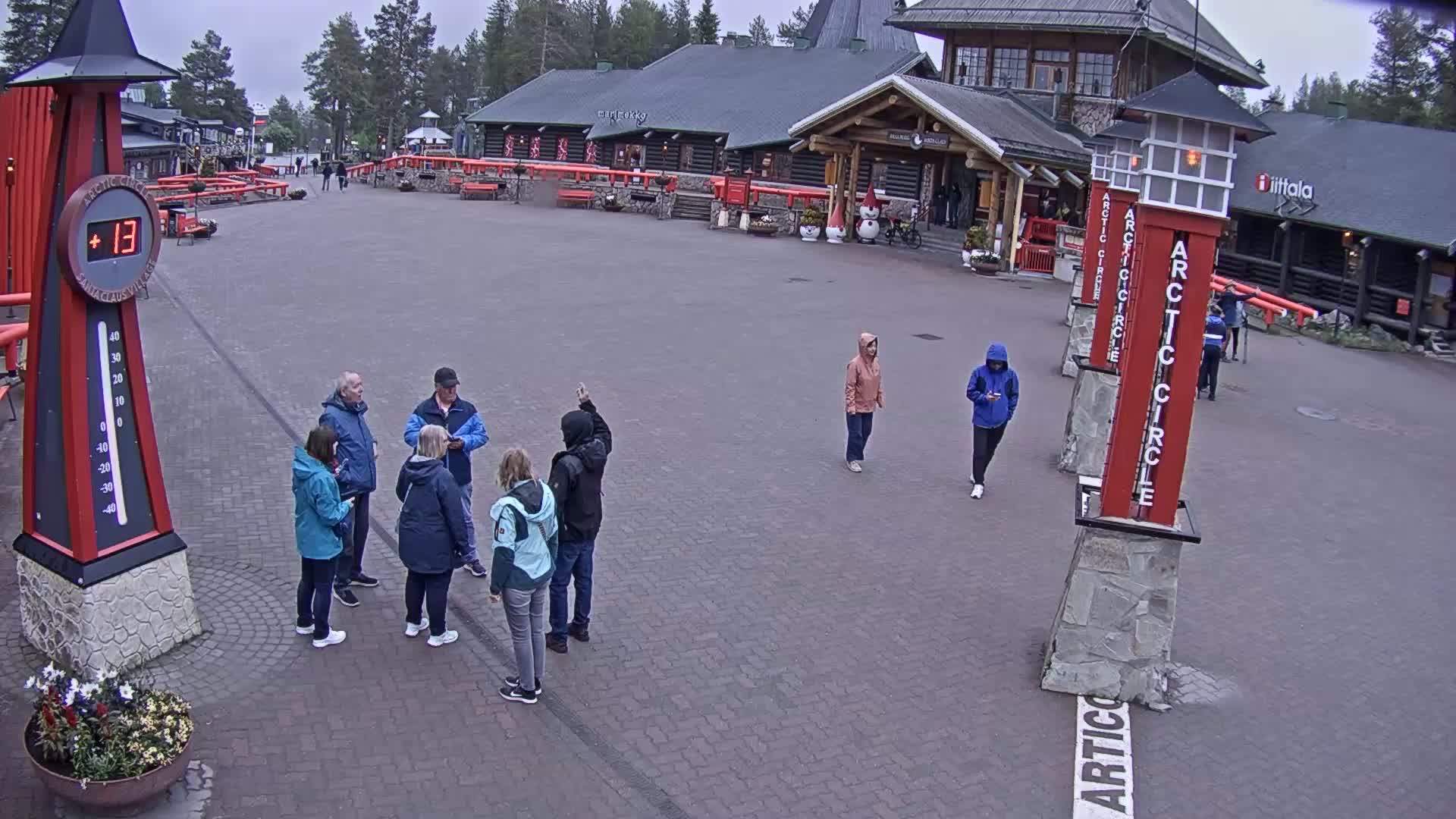 A paved plaza with several people standing in small groups, a large thermometer showing a temperature of 13 degrees, and several buildings in the background on an overcast day.