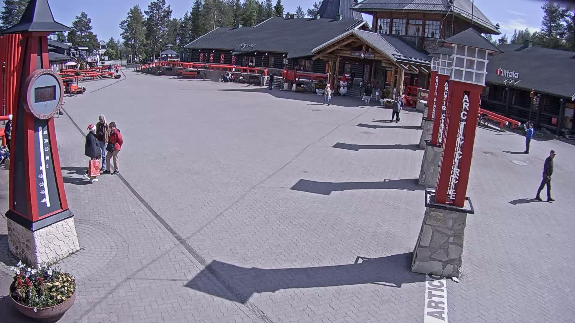 A paved plaza with several people, a large building, and tall red markers, all under sunny skies.