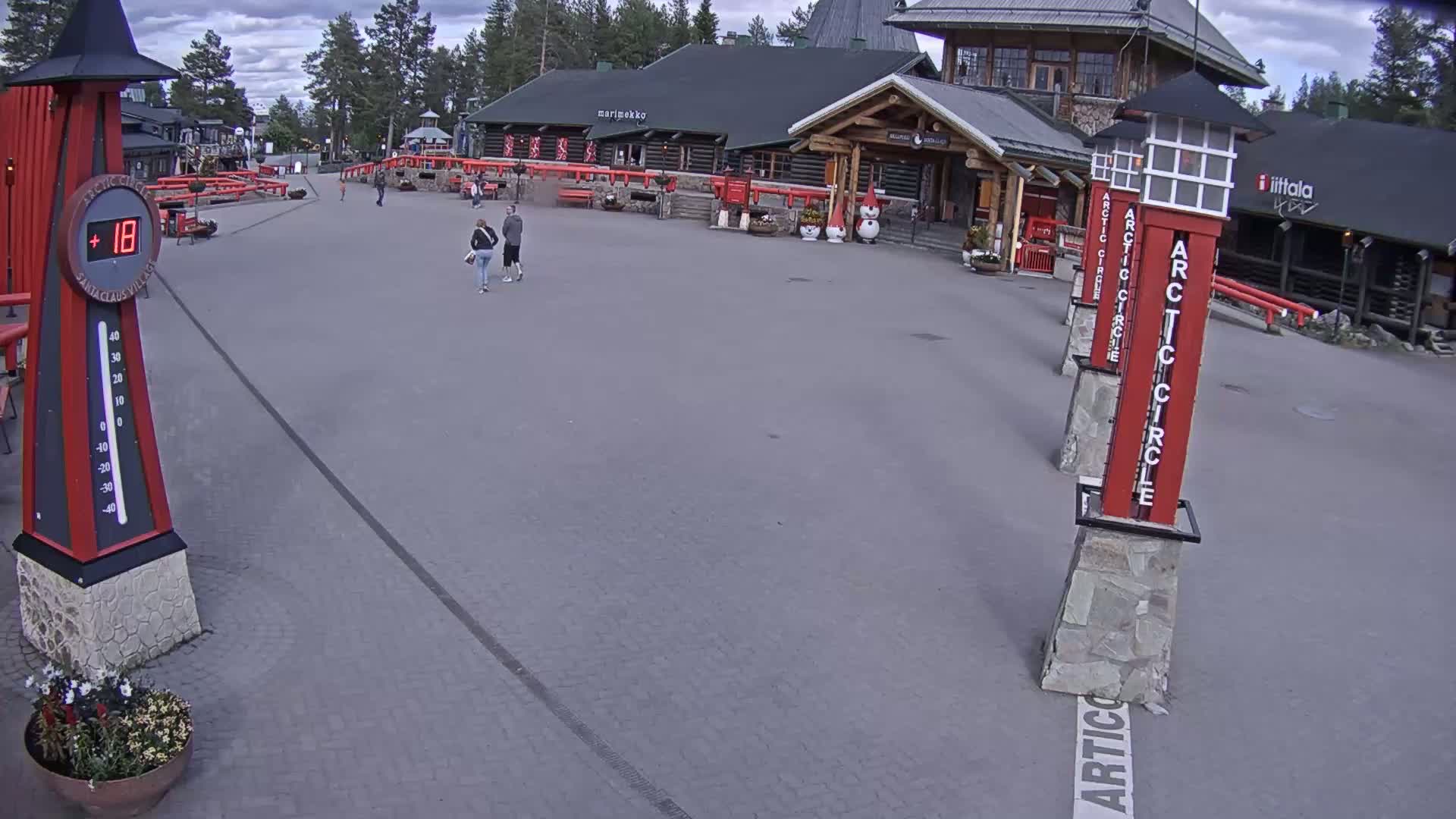 A paved plaza with several people walking near log buildings under a partly cloudy sky, featuring a large thermometer indicating a temperature of 18 degrees.