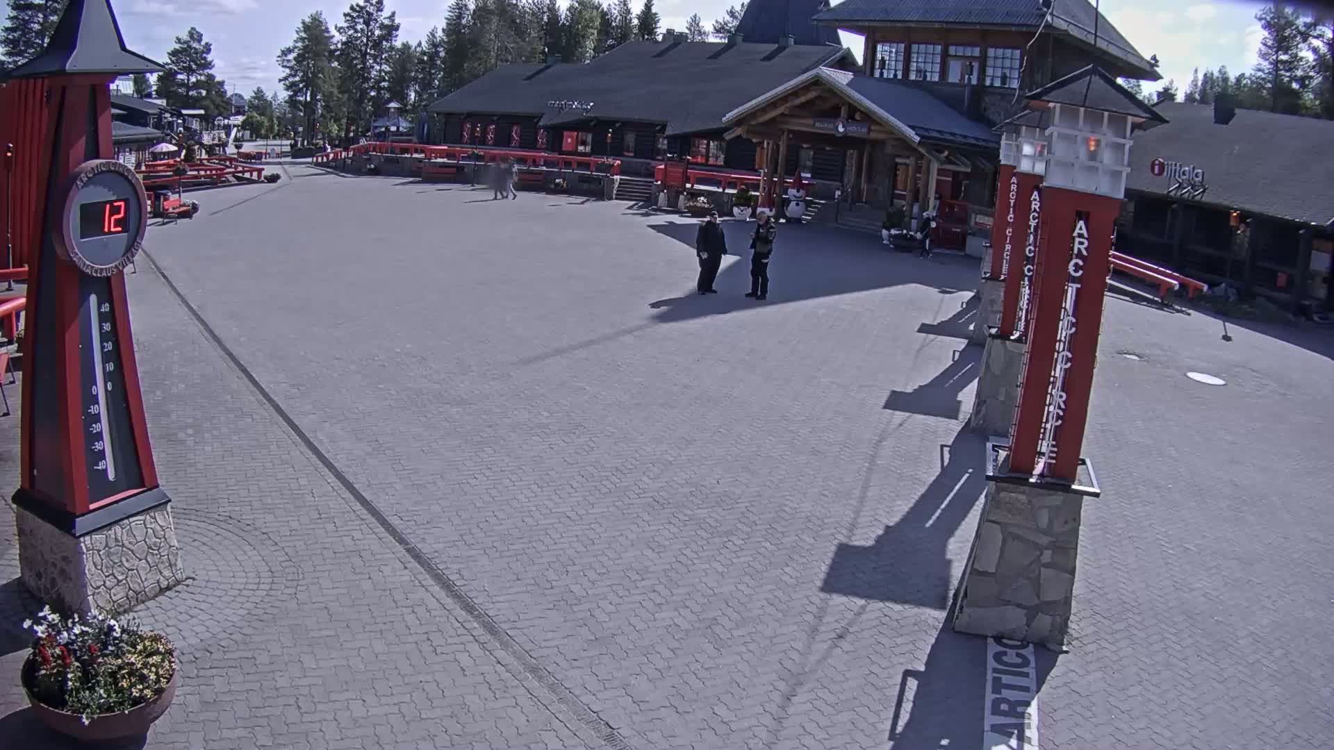 A paved plaza with a thermometer showing 12 degrees, two people standing near a building, and several tall, thin red posts.
