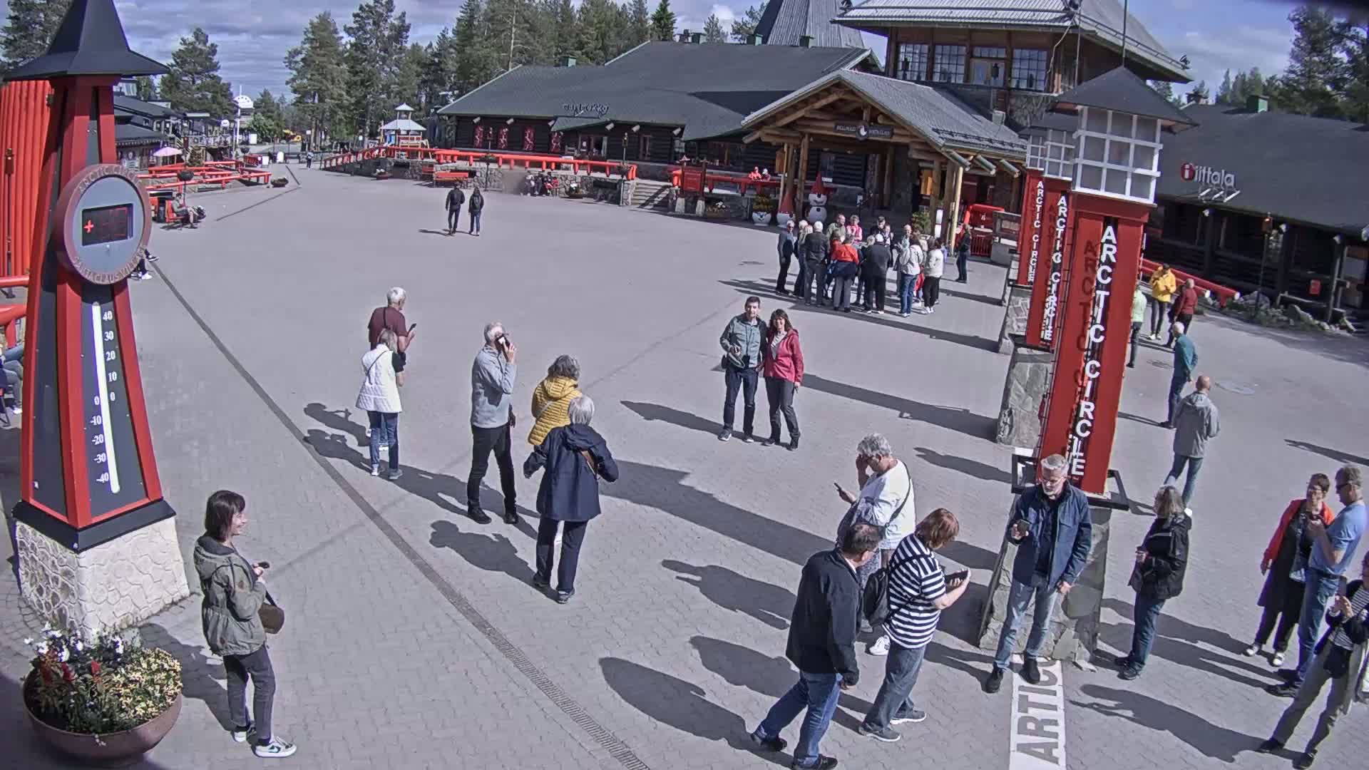 A sunny outdoor plaza with numerous people milling about near several buildings, a large thermometer, and tall red markers.