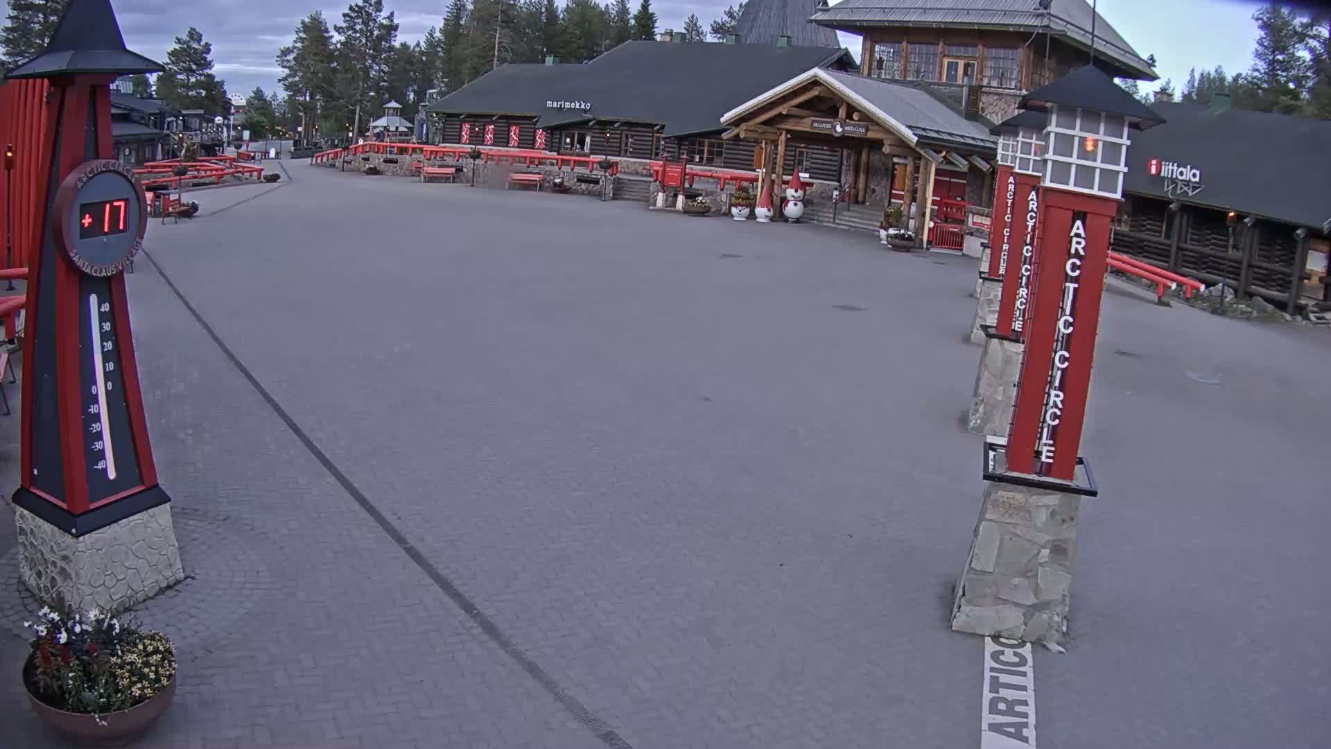 A paved plaza with log buildings and several tall, red markers indicating the Arctic Circle, under an overcast sky, shows a temperature of 17 degrees.