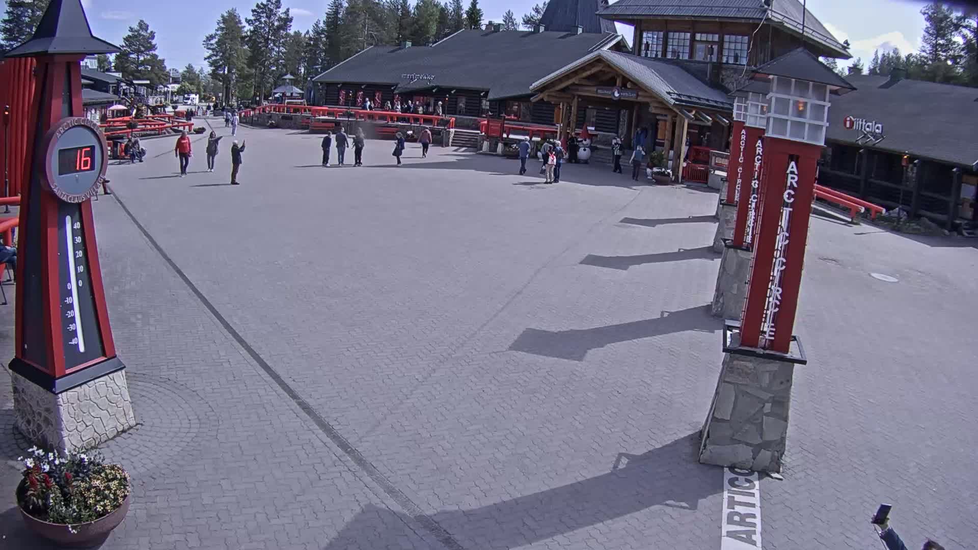 A paved plaza with several people and buildings under sunny conditions, featuring tall red pillars and a large thermometer showing a temperature of 16 degrees.