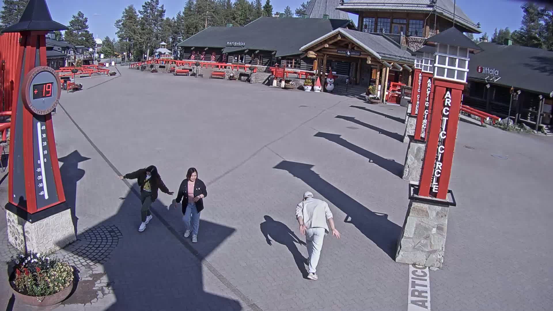 Three people walk across a paved plaza on a sunny day in front of log buildings.