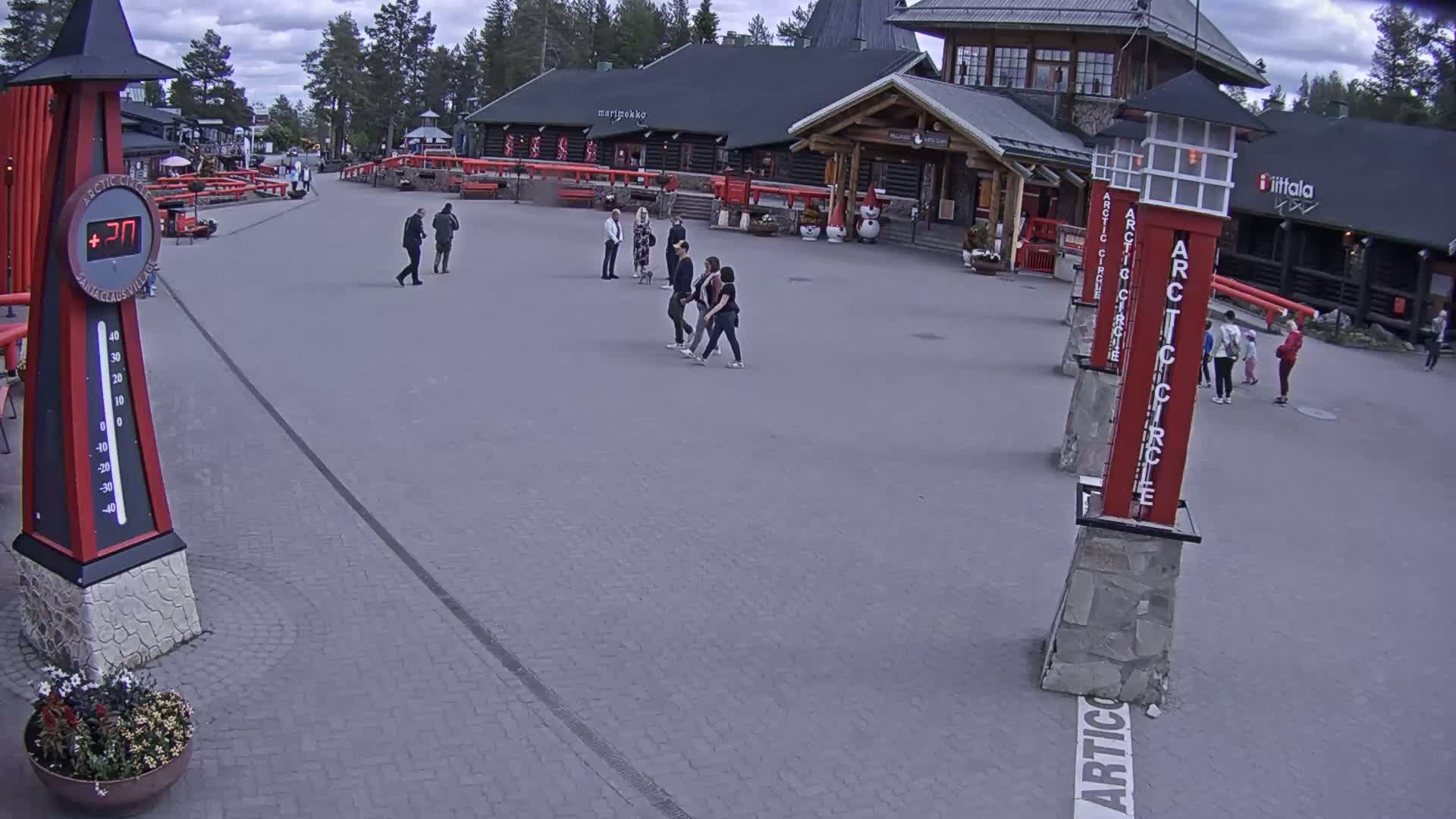 A paved plaza with several people walking, surrounded by low buildings under a partly cloudy sky.