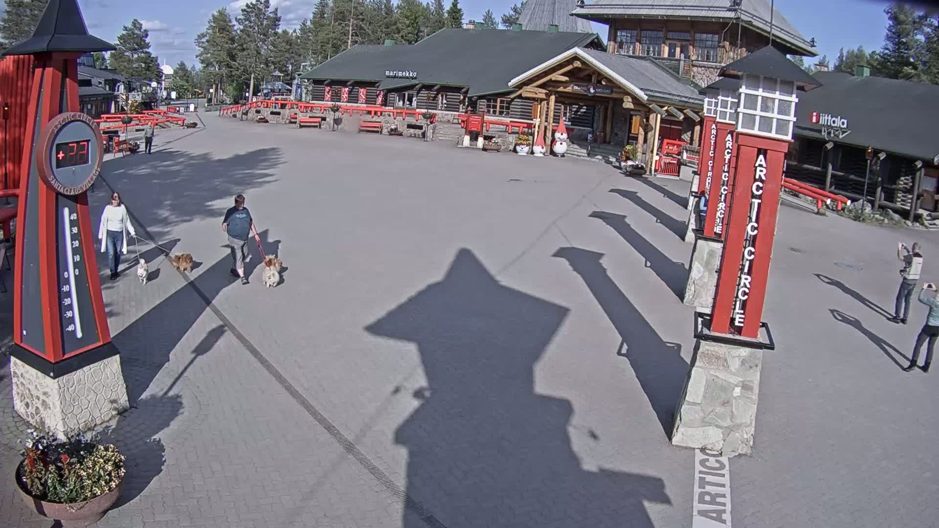 A paved plaza on a sunny day features several people walking dogs, a large thermometer, and several tall red pillars.