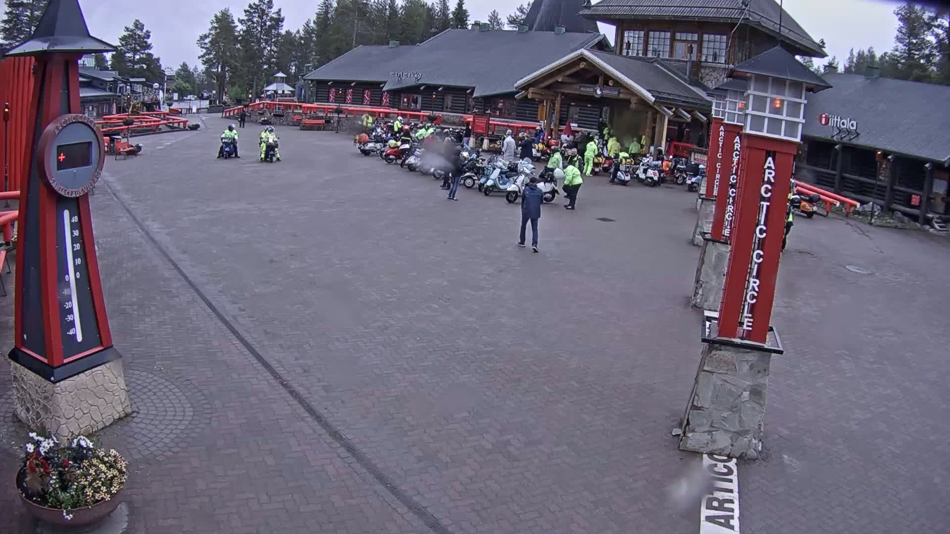 A brick plaza with several people and a group of scooters parked near a log cabin building on an overcast day.