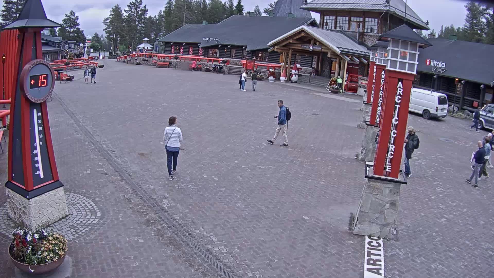 A paved plaza with several people walking, surrounded by wooden buildings under an overcast sky.