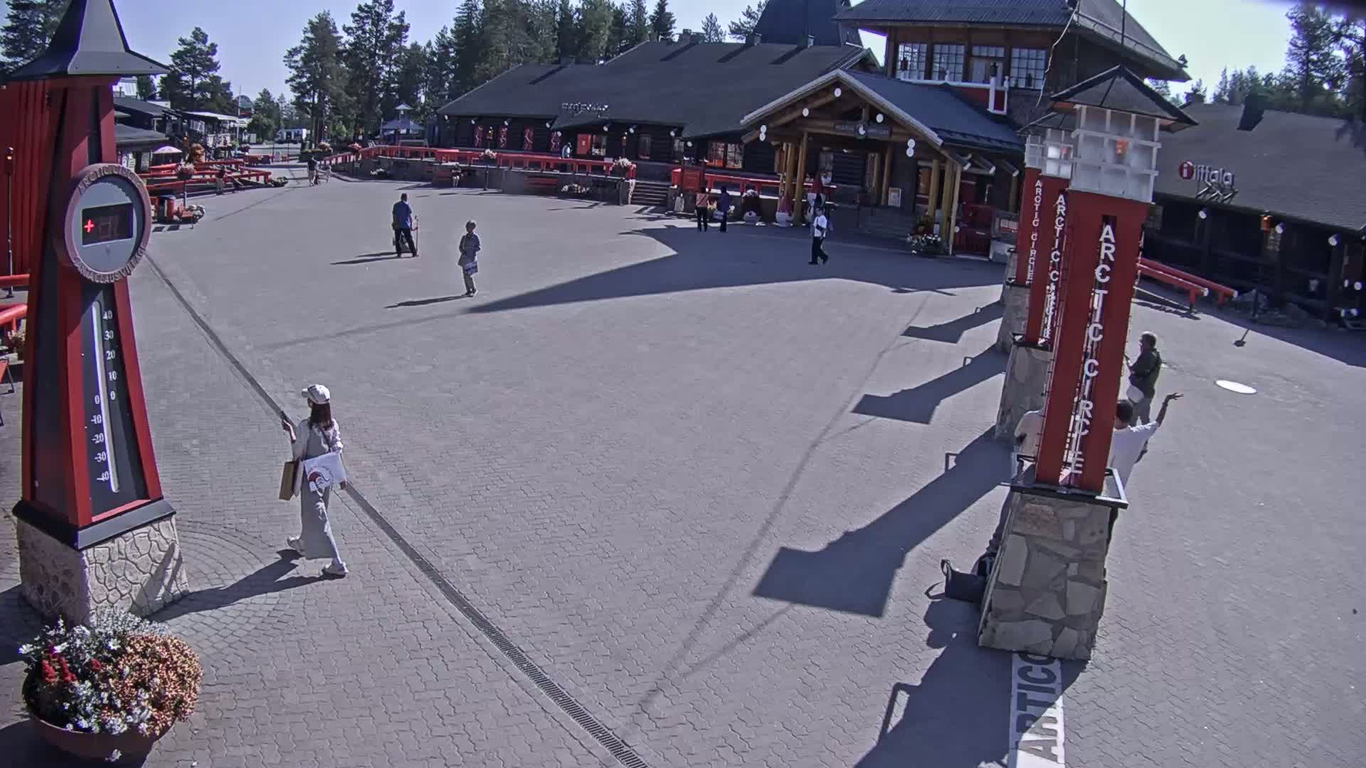 A paved plaza with a few people walking, near a building complex on a sunny day.